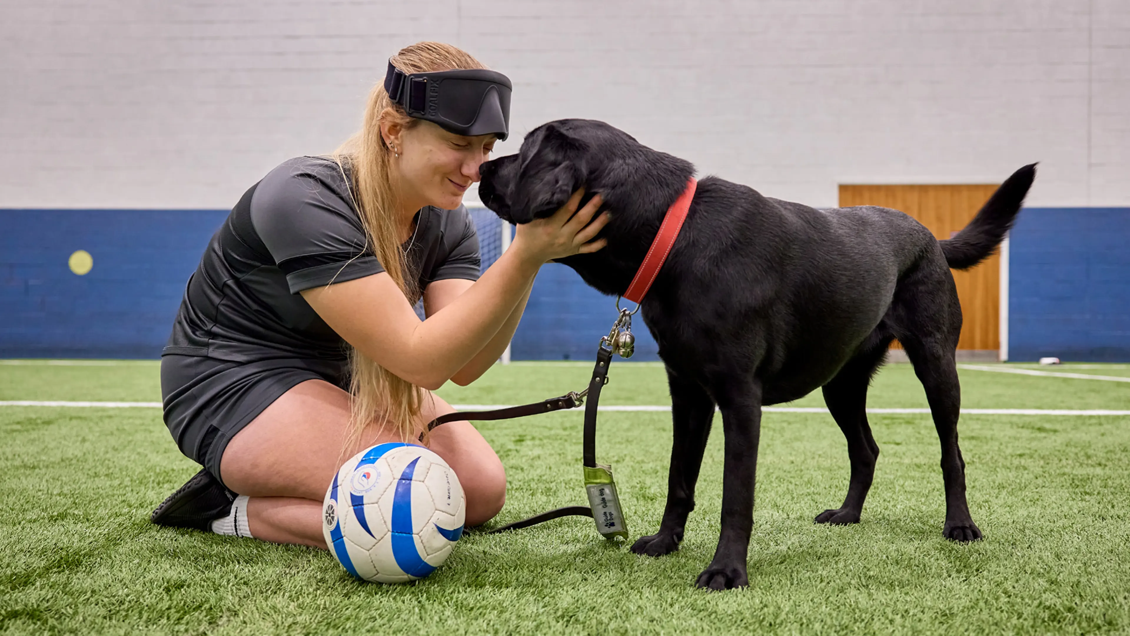Guide dog owner Sam in sports gear with a football next to her gently touching black Labrador Helga's face. 