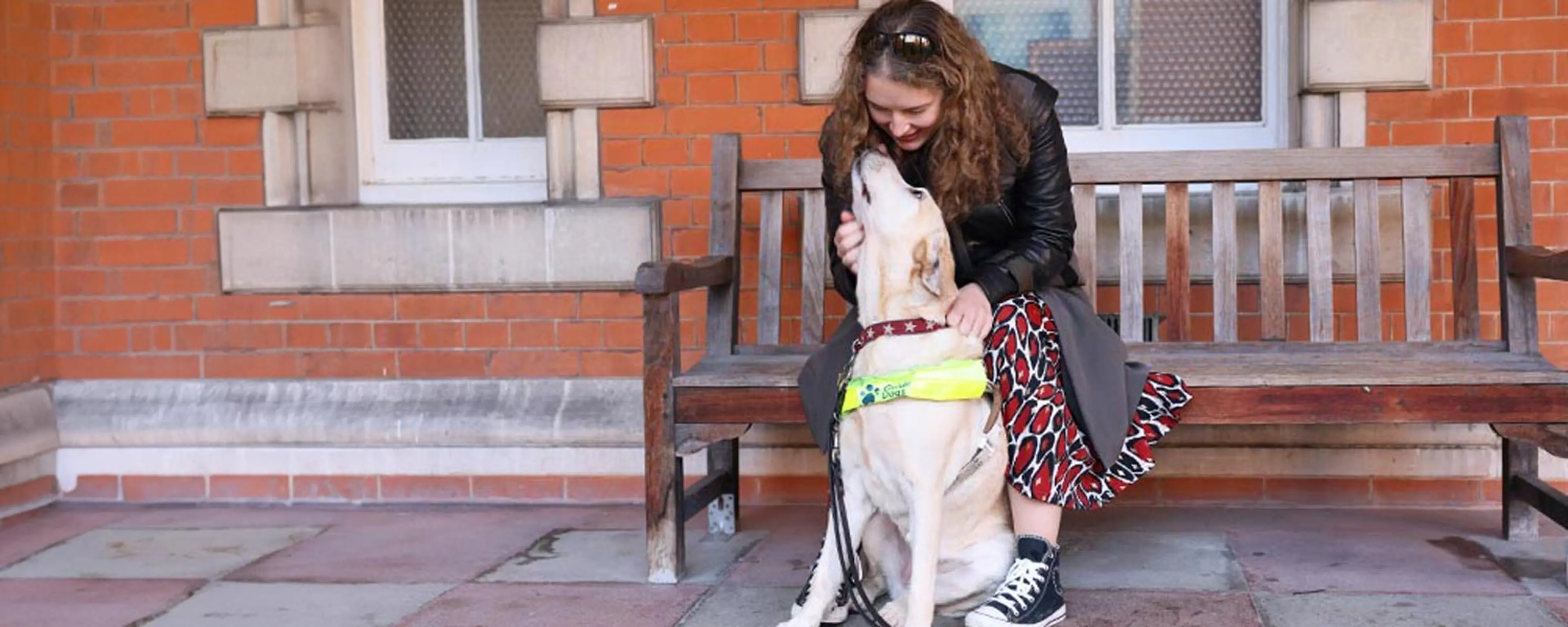 Guide dog owner Anica sitting on a bench looking down at Labrador guide dog Lassie who is looking up towards her.