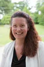 Kate smiles at the camera. She has long, curly, dark red hair and is wearing a black and white top.