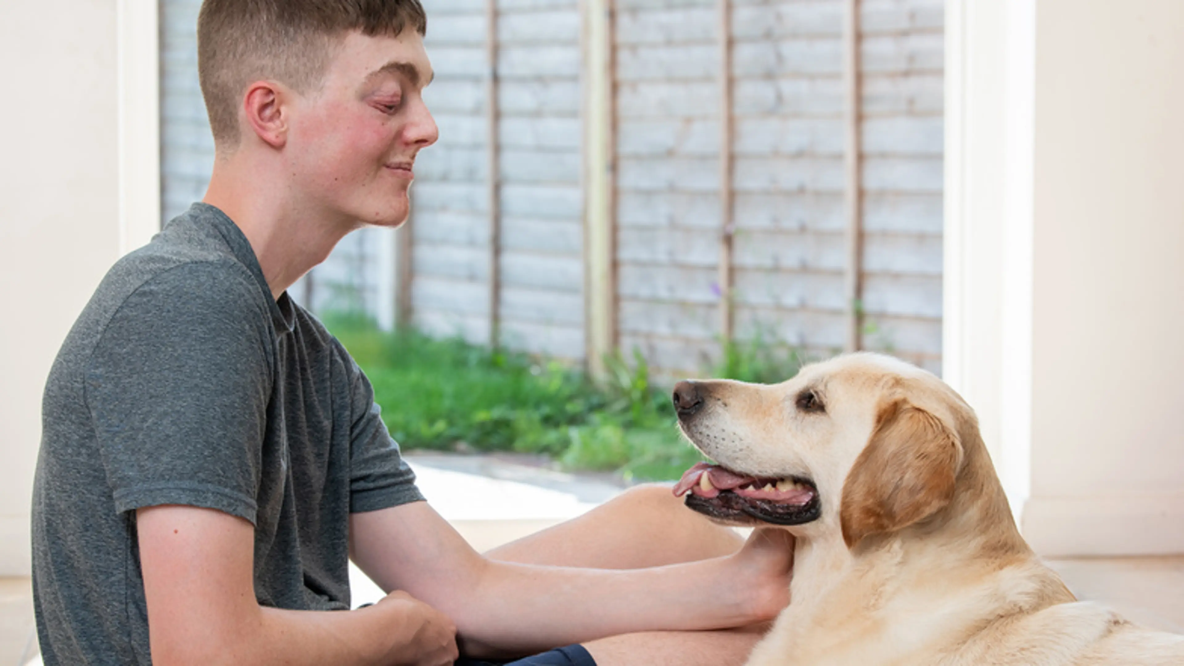 Declan sits on the floor of his home smiling at his buddy dog Barney, a yellow Labrador who looks back at him.