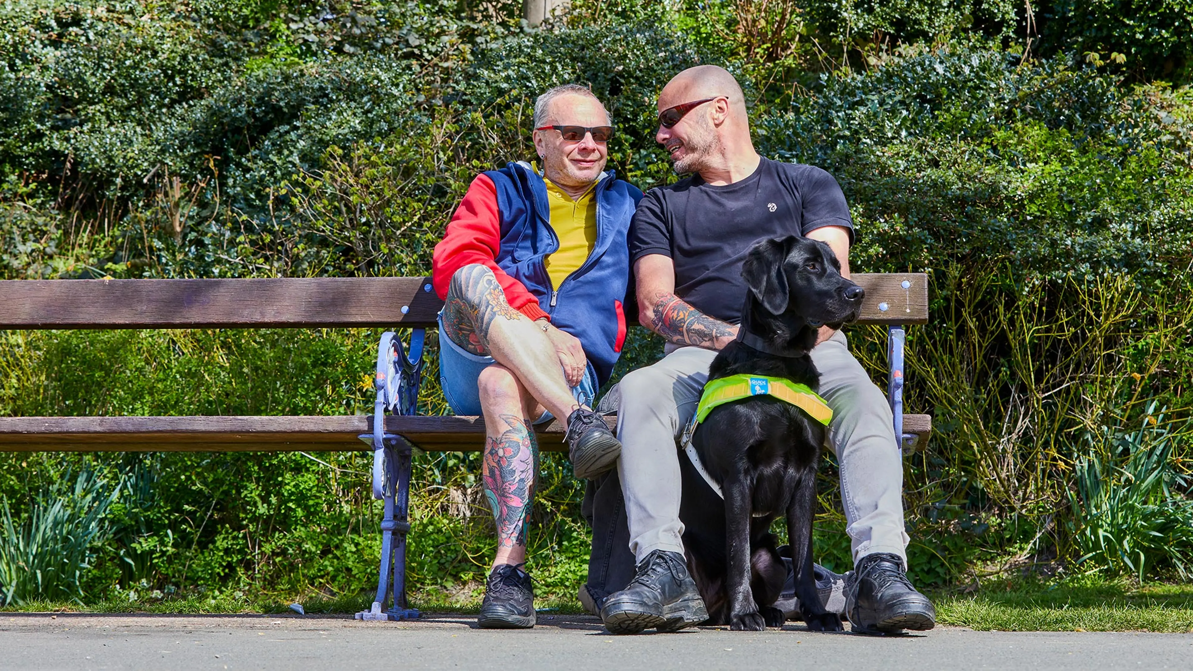 Guide dog owner Terry with his guide dog Spencer, sitting next to another man on a bench and both are smiling.  