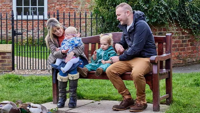 Margot, her parents, and her sister, sit on a bench looking at some ducks.