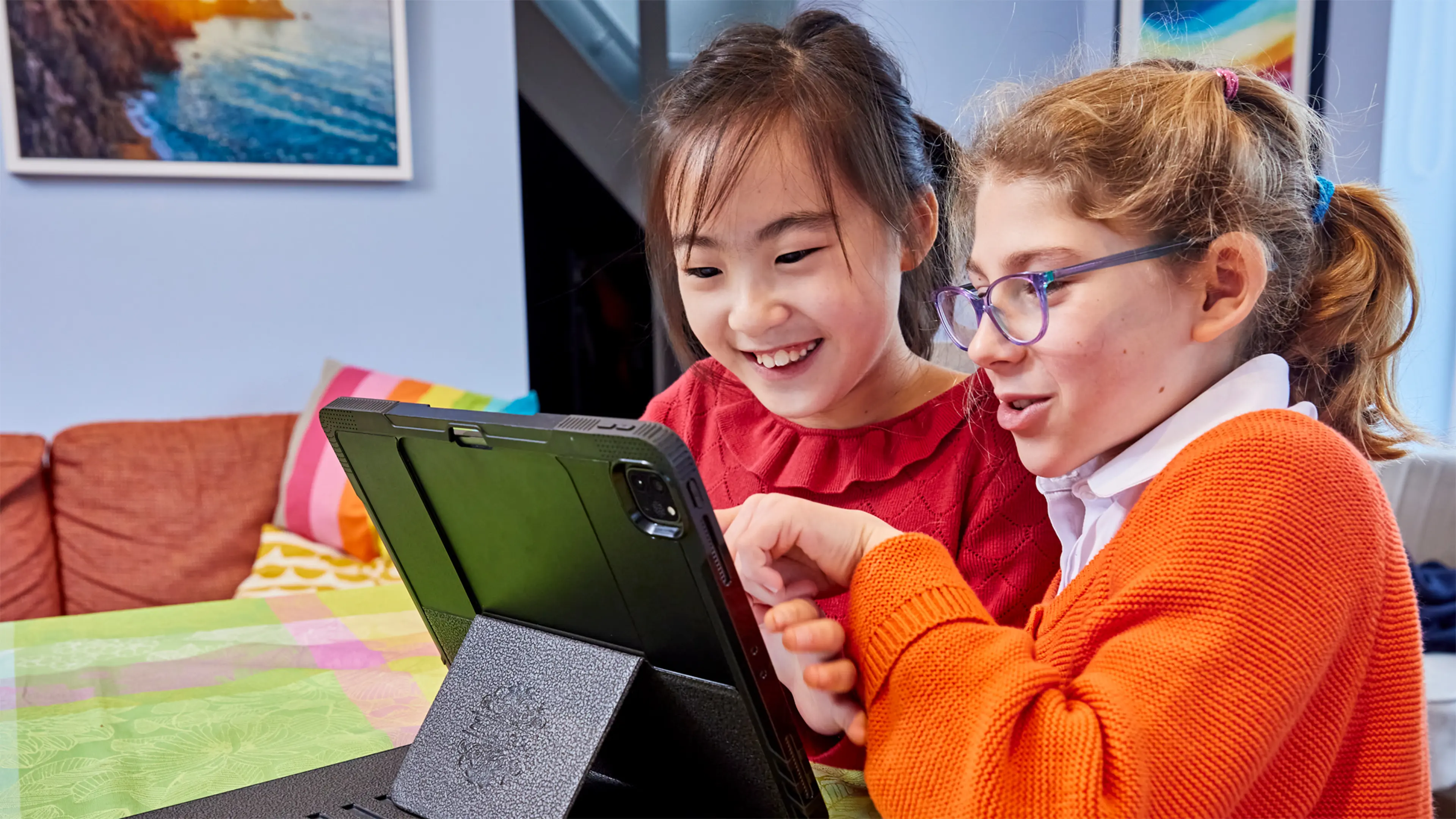 Josie, a child who has a vision impairment, sits at a table with her friend. They are looking at the screen of a piece of assistive technology.
