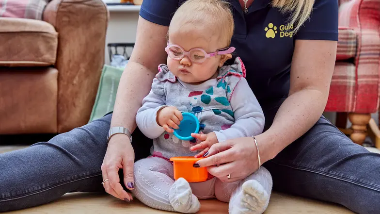 A habilitation specialist sits on the floor with a baby girl with vision impairment, playing with colourful plastic blocks.
