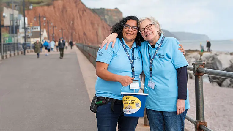 Two women in Guide Dogs t-shirts stand in front of some cliffs in Devon with their arms round each other smiling. One of them holds a fundraising bucket.