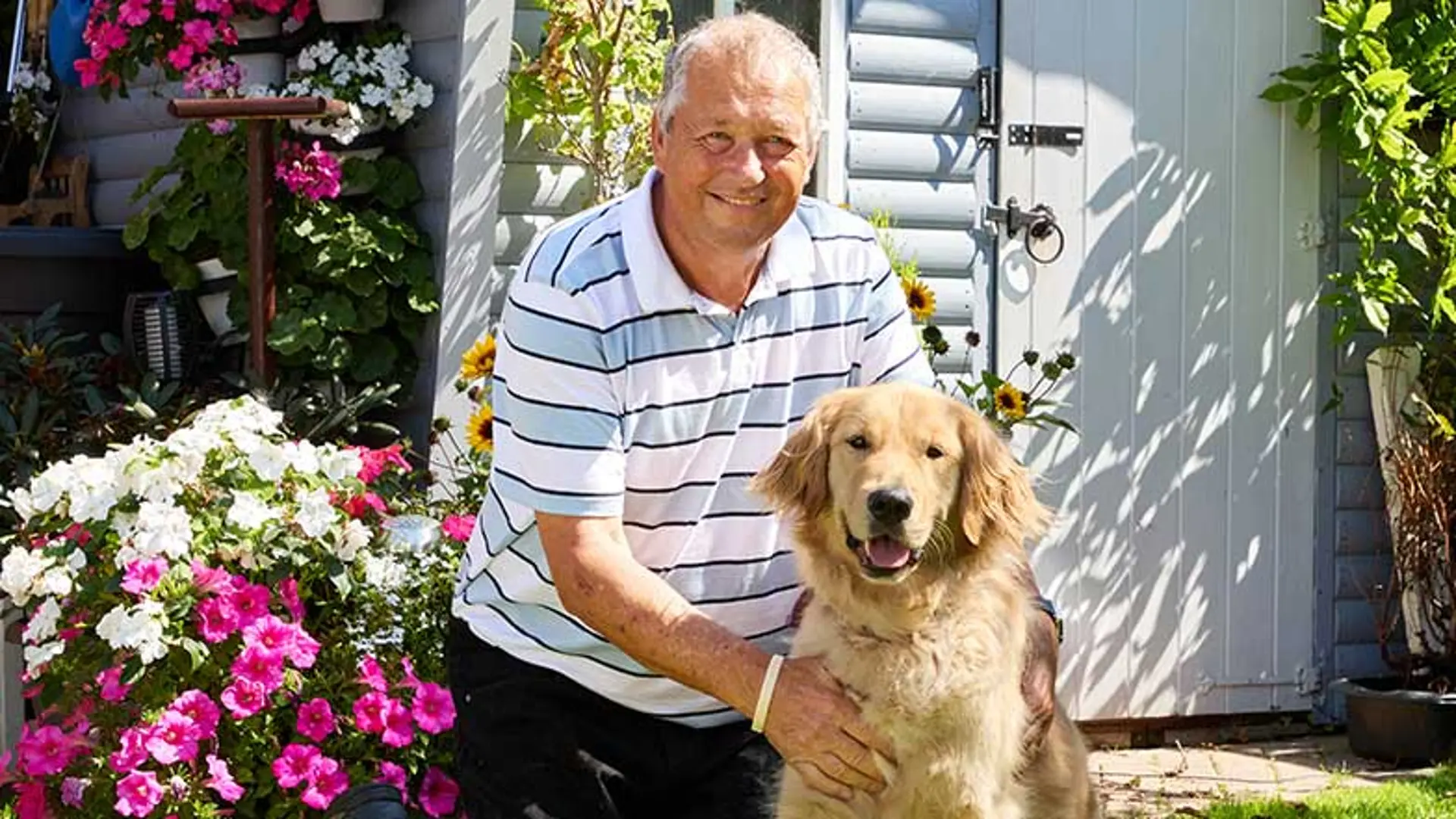 Guide dog owner Tony kneeling next to his guide dog Angus in a sunny garden with brightly coloured flowers.