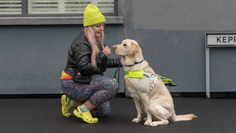 A guide dog owner kneels beside her guide dog, smiling.