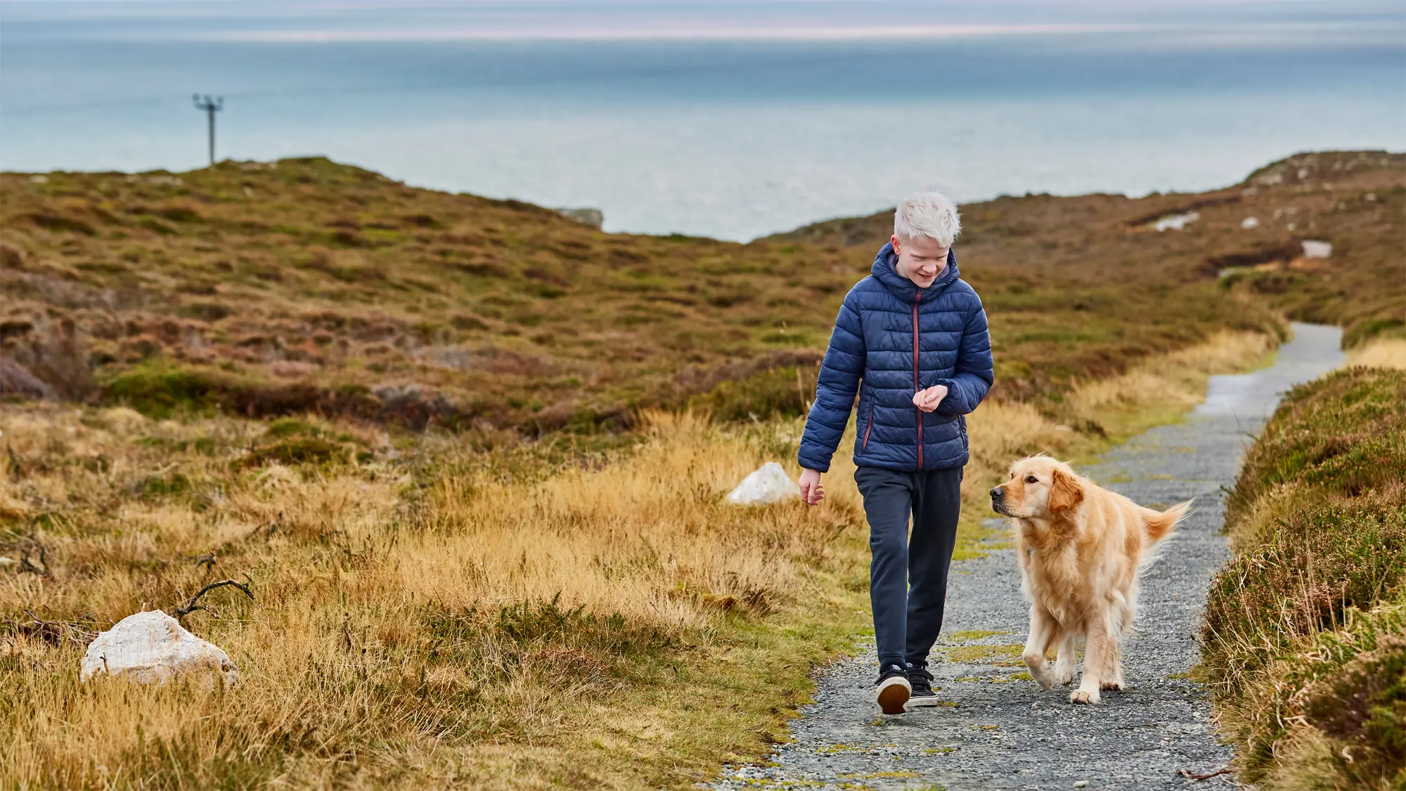 Ellis, who has albinism, walks with his buddy dog Ralph down a path, the sea is behind them.