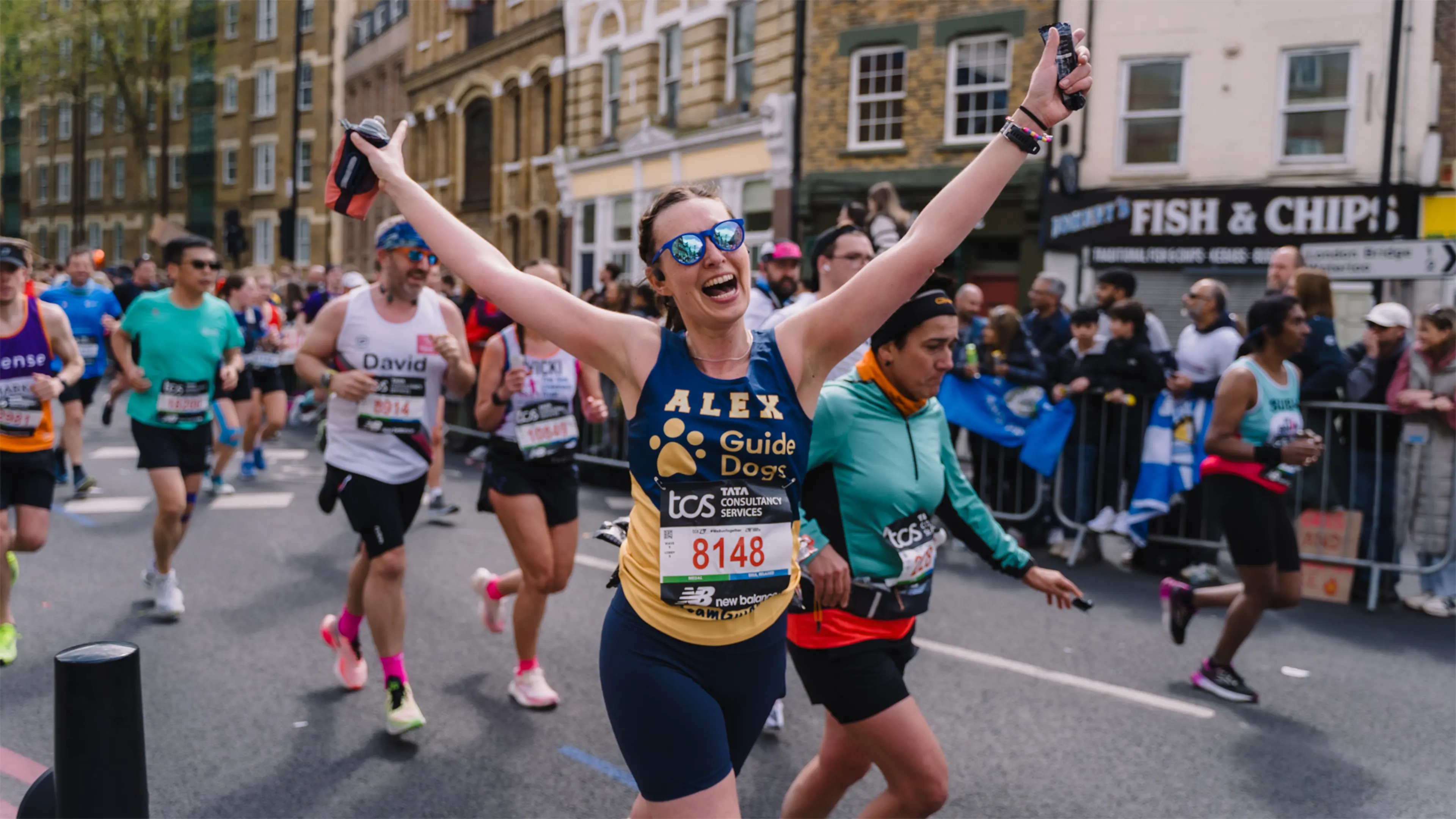 A woman in a Guide Dogs vest runs down a street during the London Marathon, her arms are raised high in the air.