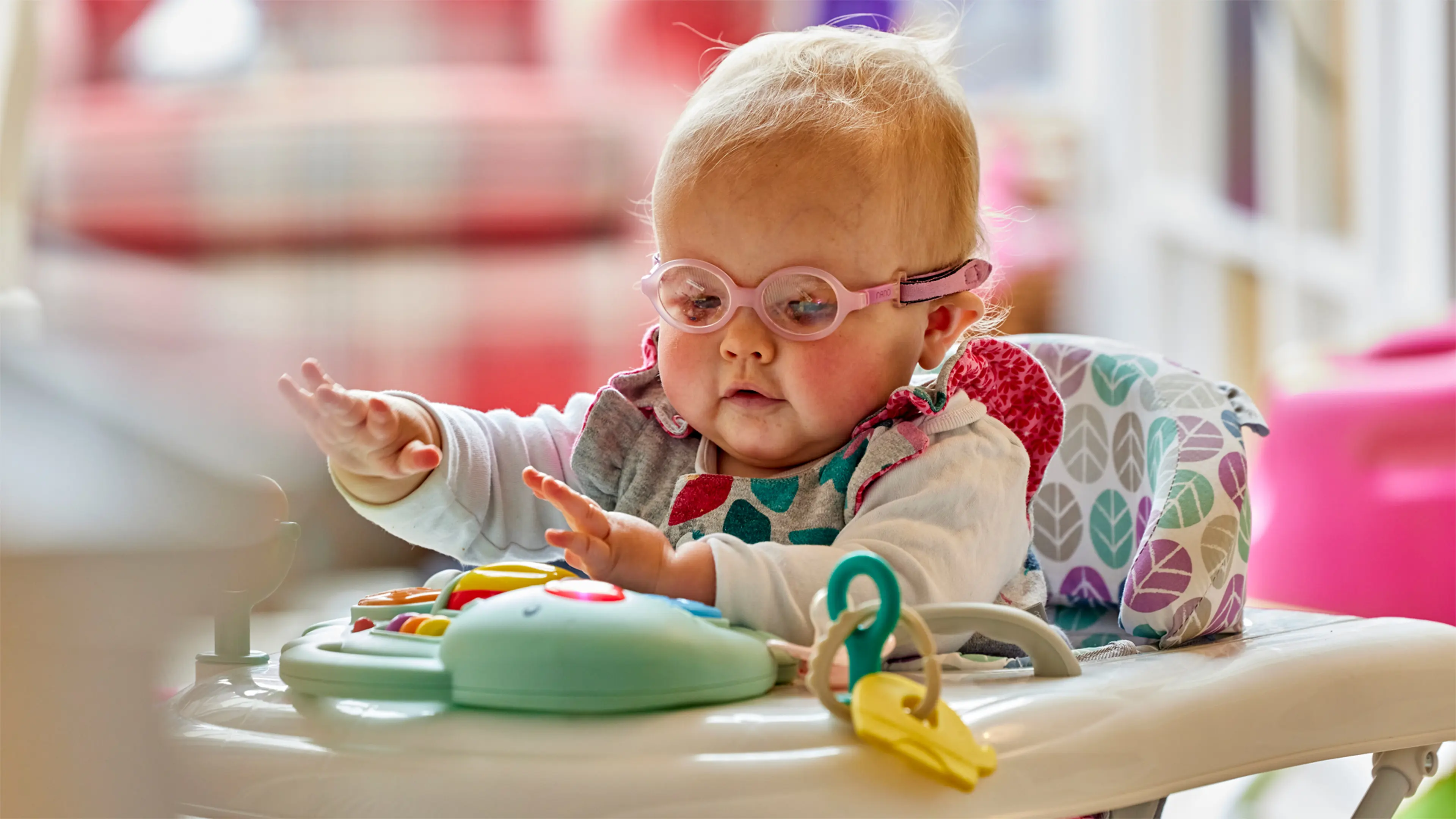 Margot, a baby who has a vision impairment, sits in a chair and plays with the sensory toys on the tray in front of her.