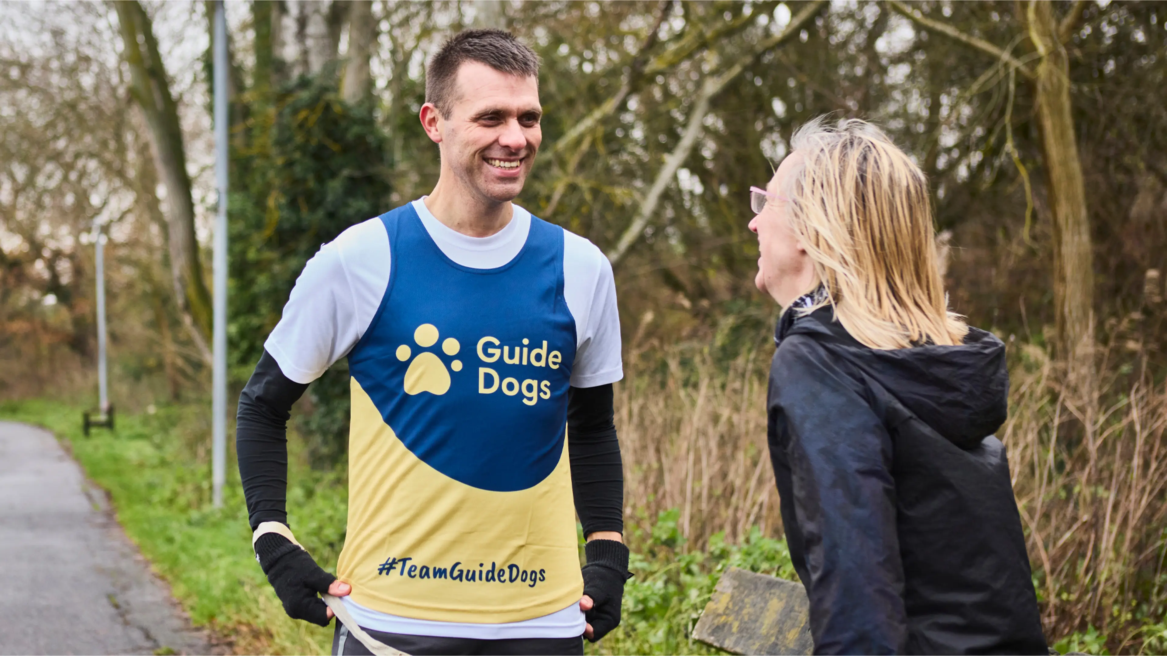 Steve, who has a vision impairment, wears a Guide Dogs running vest. He laughs in the park with his guide runner. 
