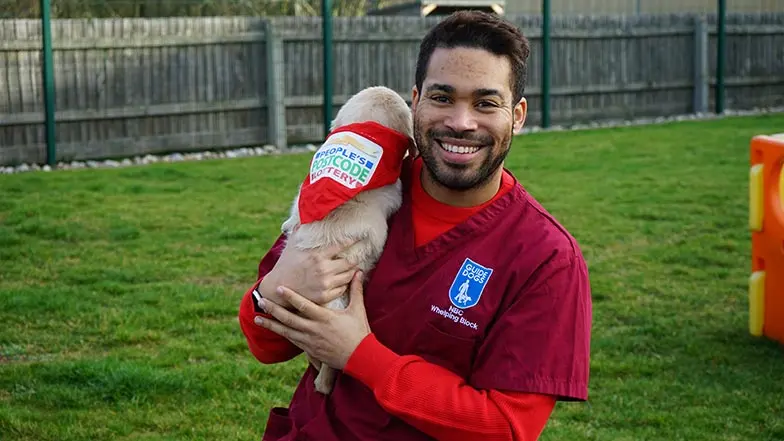 Singer, Danyl Johnson  looks to camera. He is holding a guide dog puppy who is wearing a People's Postcode Lottery bandana