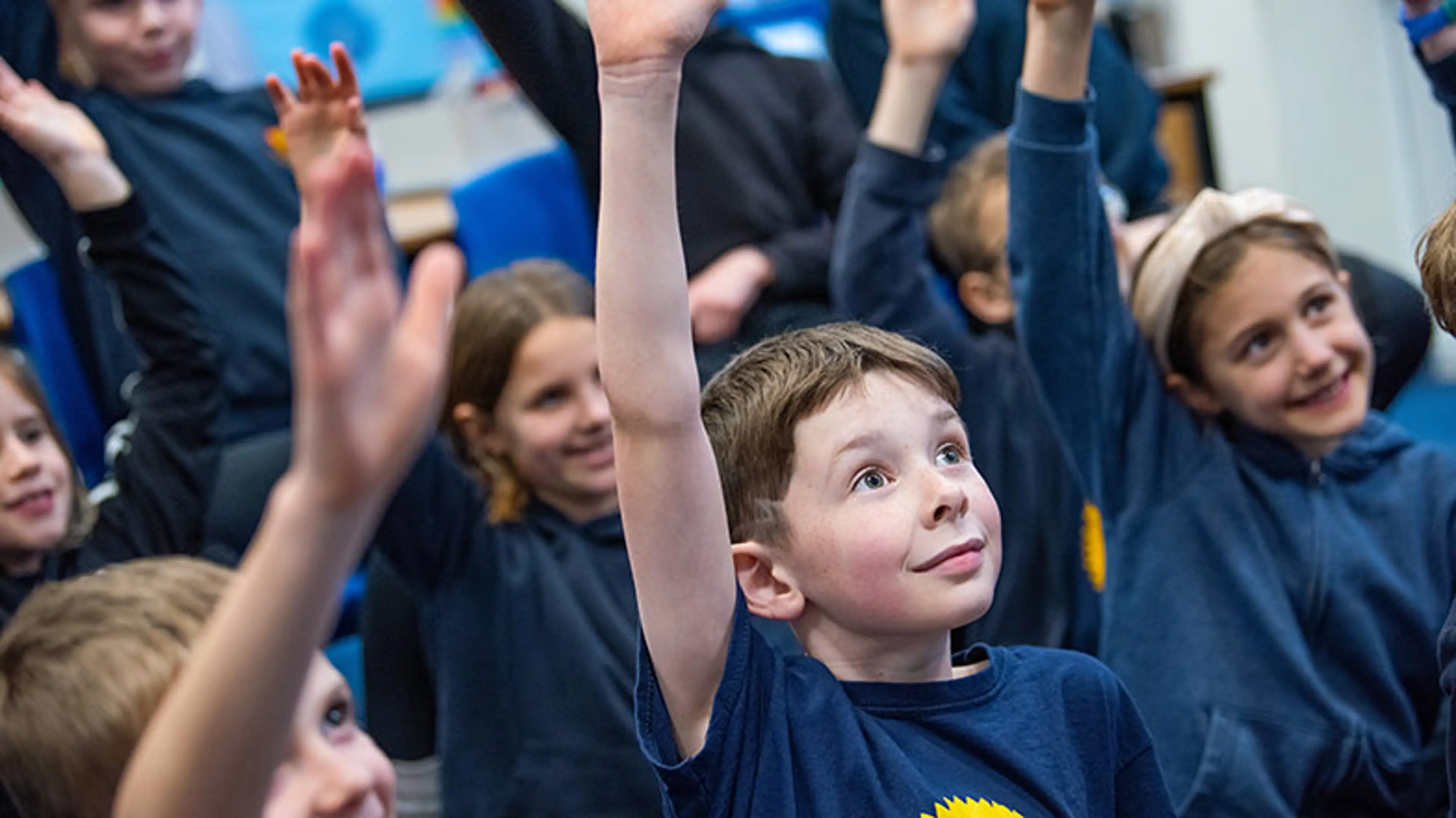 School children sitting on a classroom floor with their hands raised.