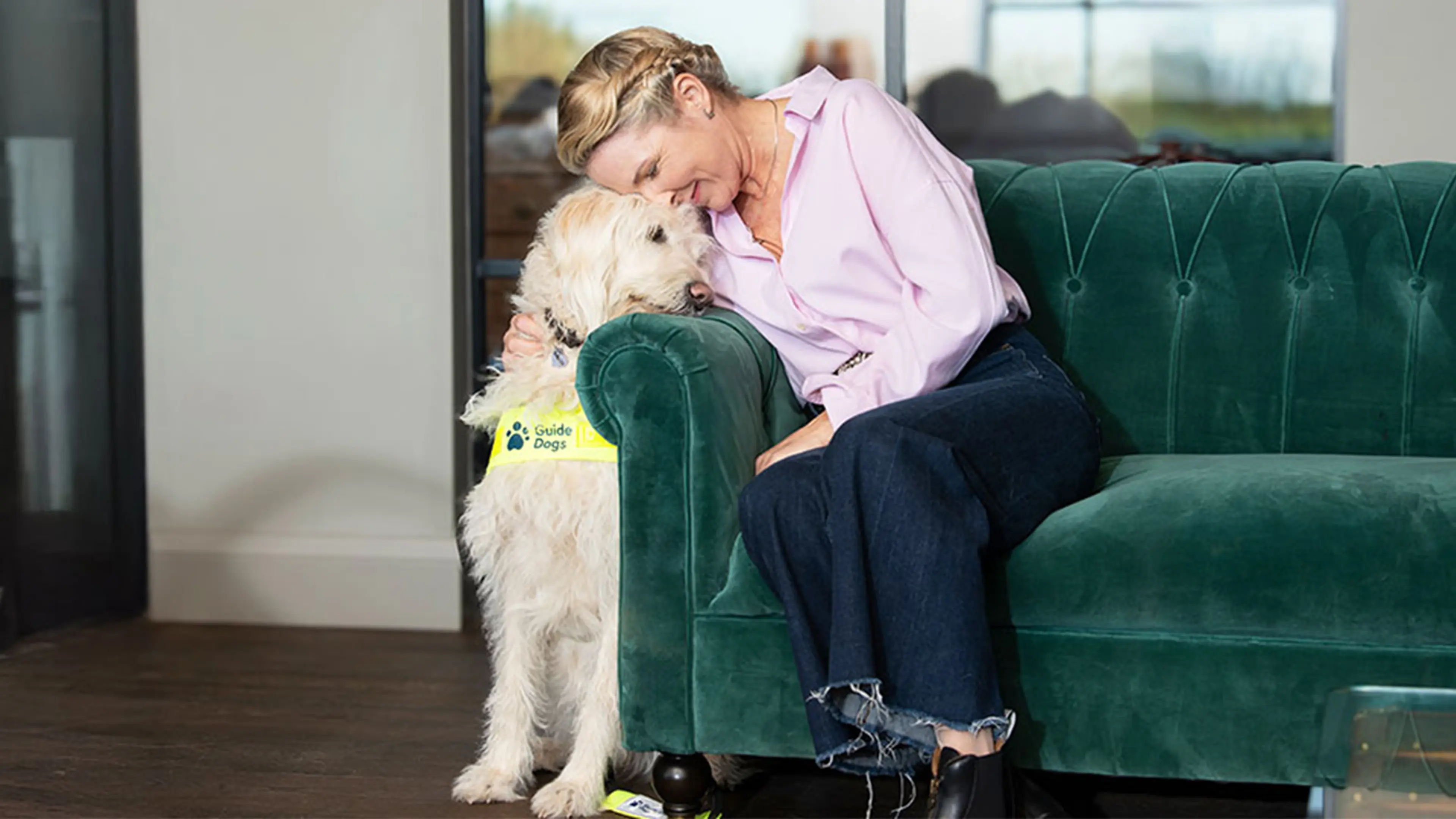 Guide dog owner Verity sitting on a couch with her head gently leaning on the top of guide dog Luna's head. Luna is gently resting her chin on the arm of the couch.