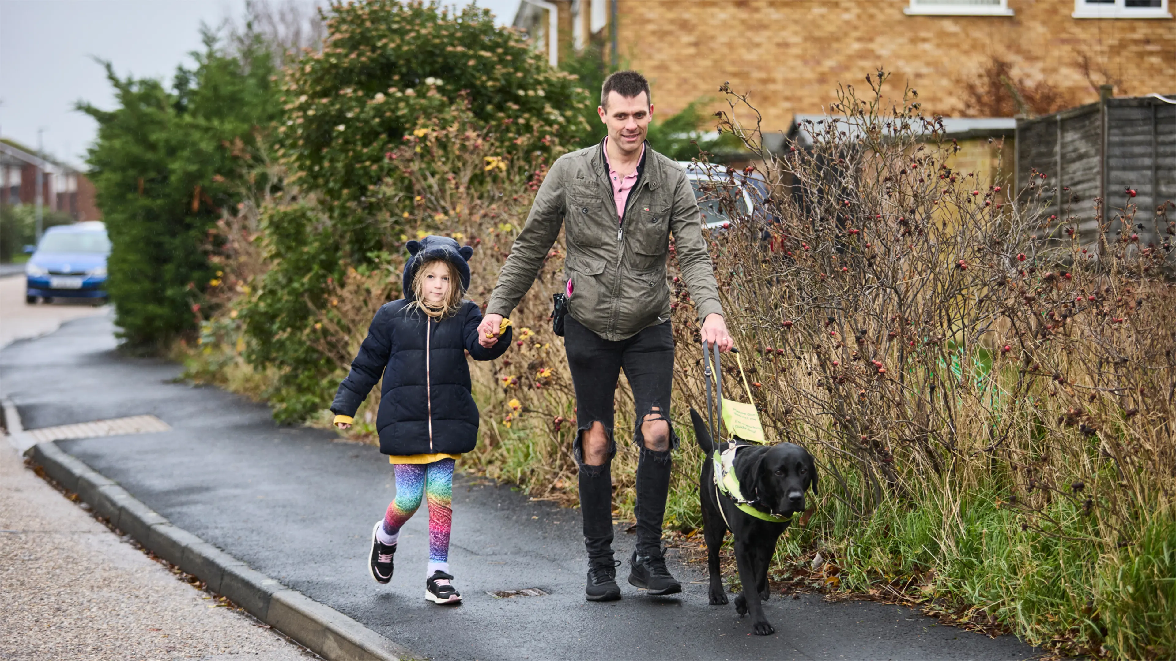 Guide dog owner Steve walks along the pavement holding his daughter's hand, with his guide dog Wilf.