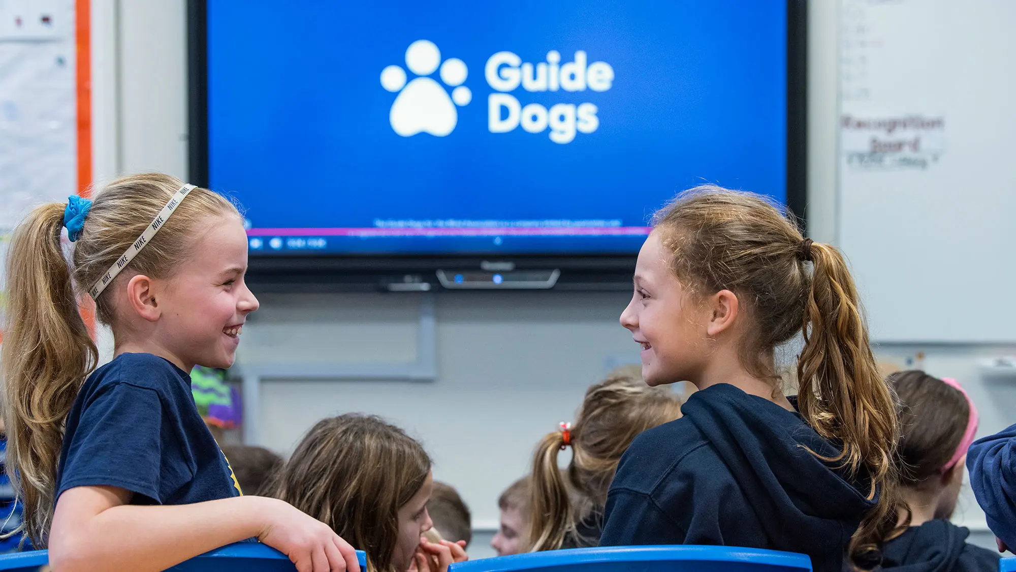 Two schoolgirls in a classroom smiling at each other with the board showing Guide Dogs logo in the background.