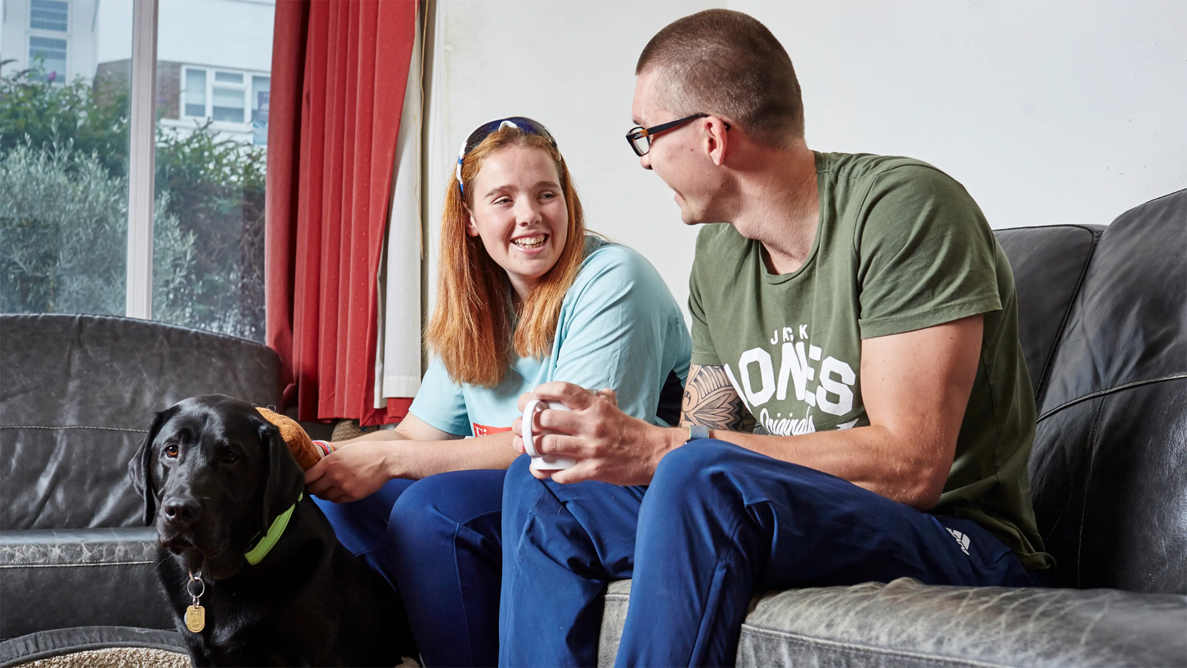 Lois, a guide dog owner, sits on the sofa with her partner. At her feet is her guide dog, black Labrador Buster.