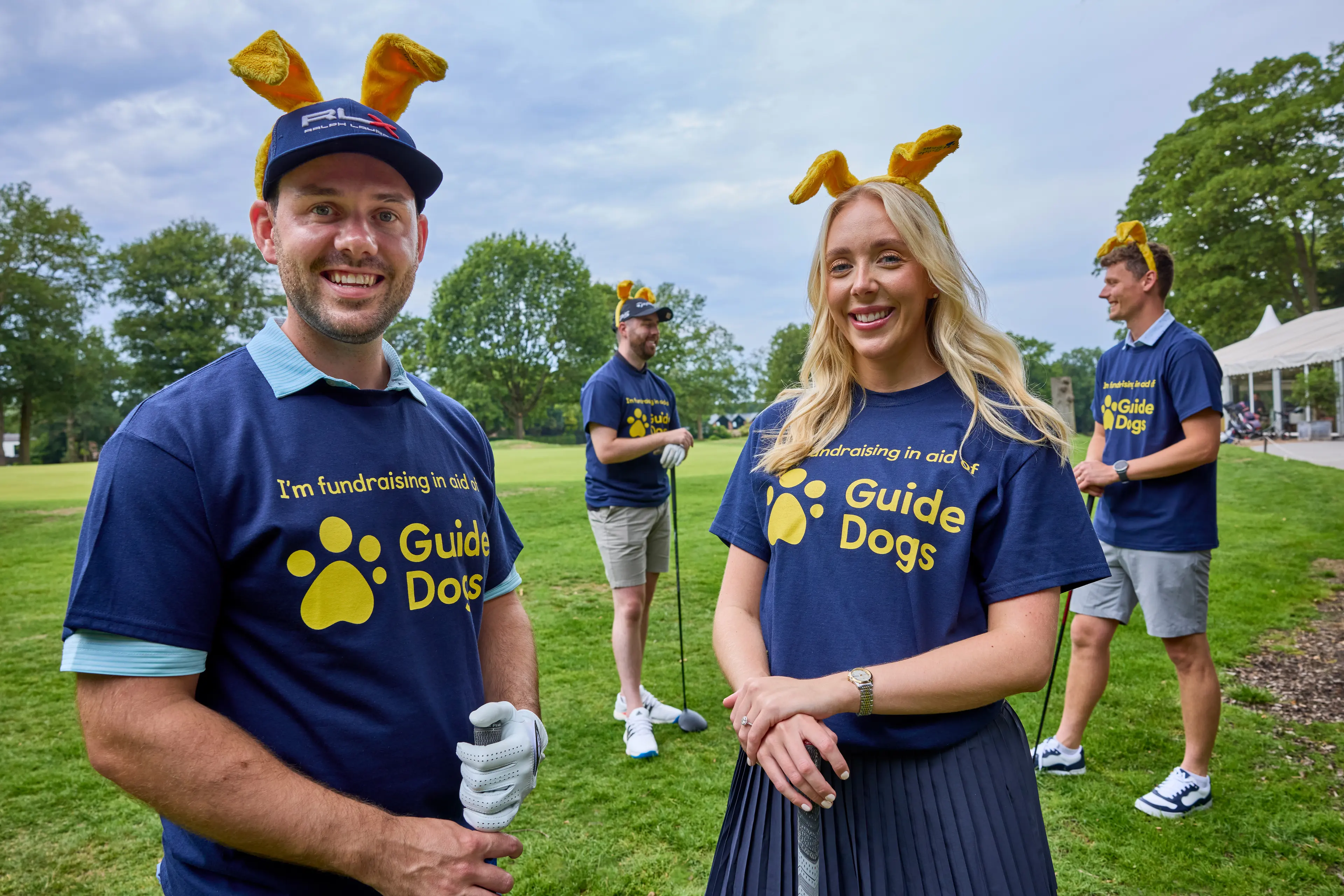A woman and 2 men stand on a golf course wearing Guide Dogs t-shirts and ears.