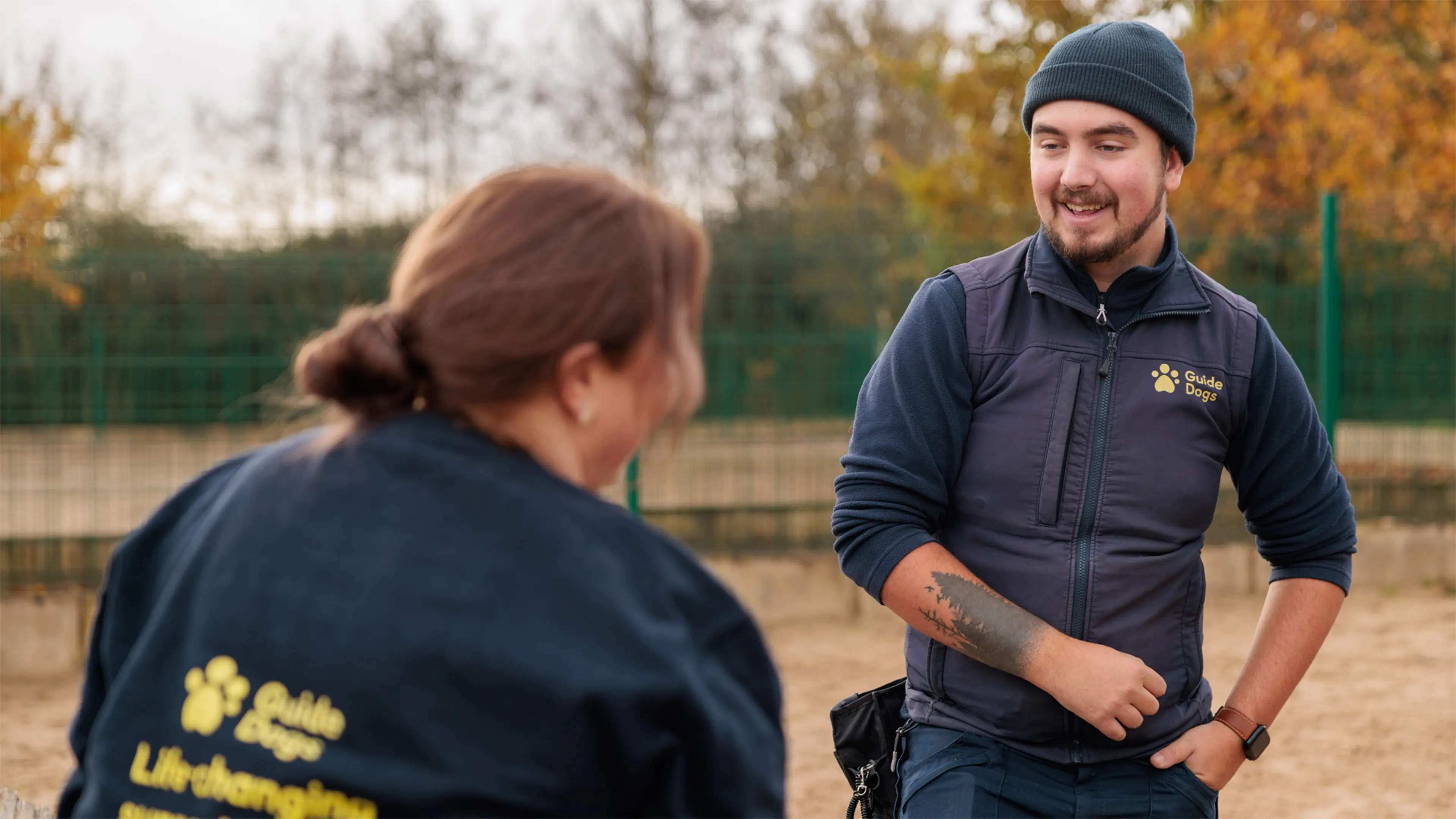 A Guide Dog Trainer stands outside in a training area talking to a colleague. He wears a Guide Dogs gilet and a treat pouch.