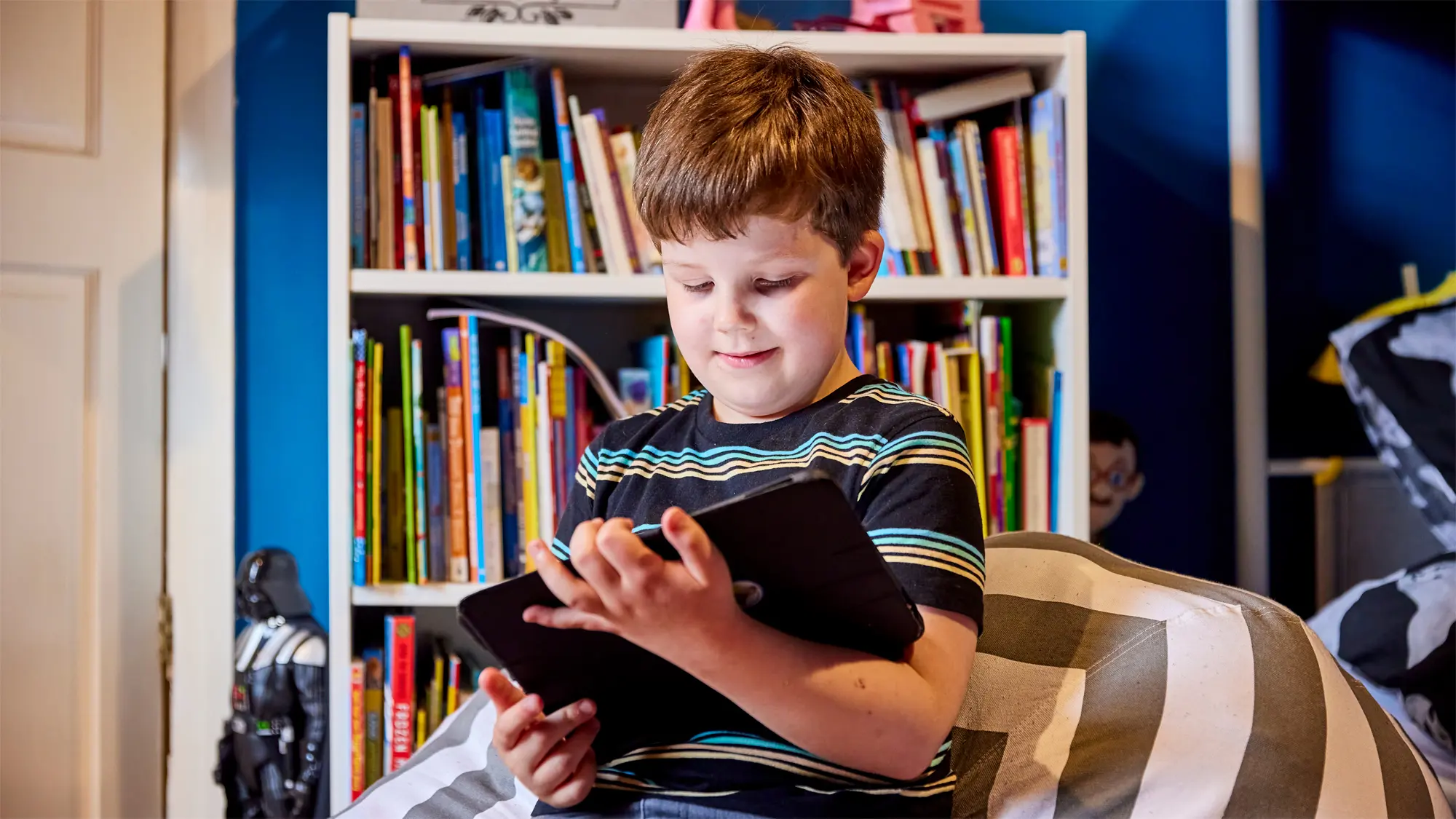 A young boy with a vision impairment sits on a beanbag in his bedroom using his iPad. 