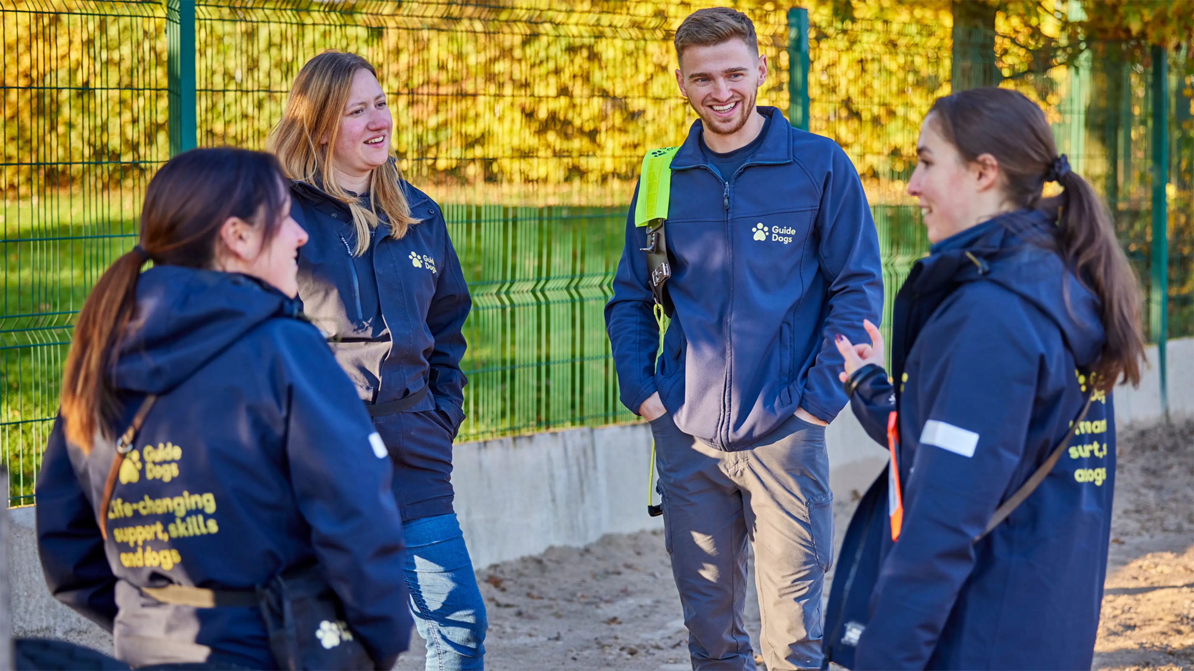 Four Guide Dogs Academy staff members stand in a circle outside smiling and talking in their Guide Dogs uniform.