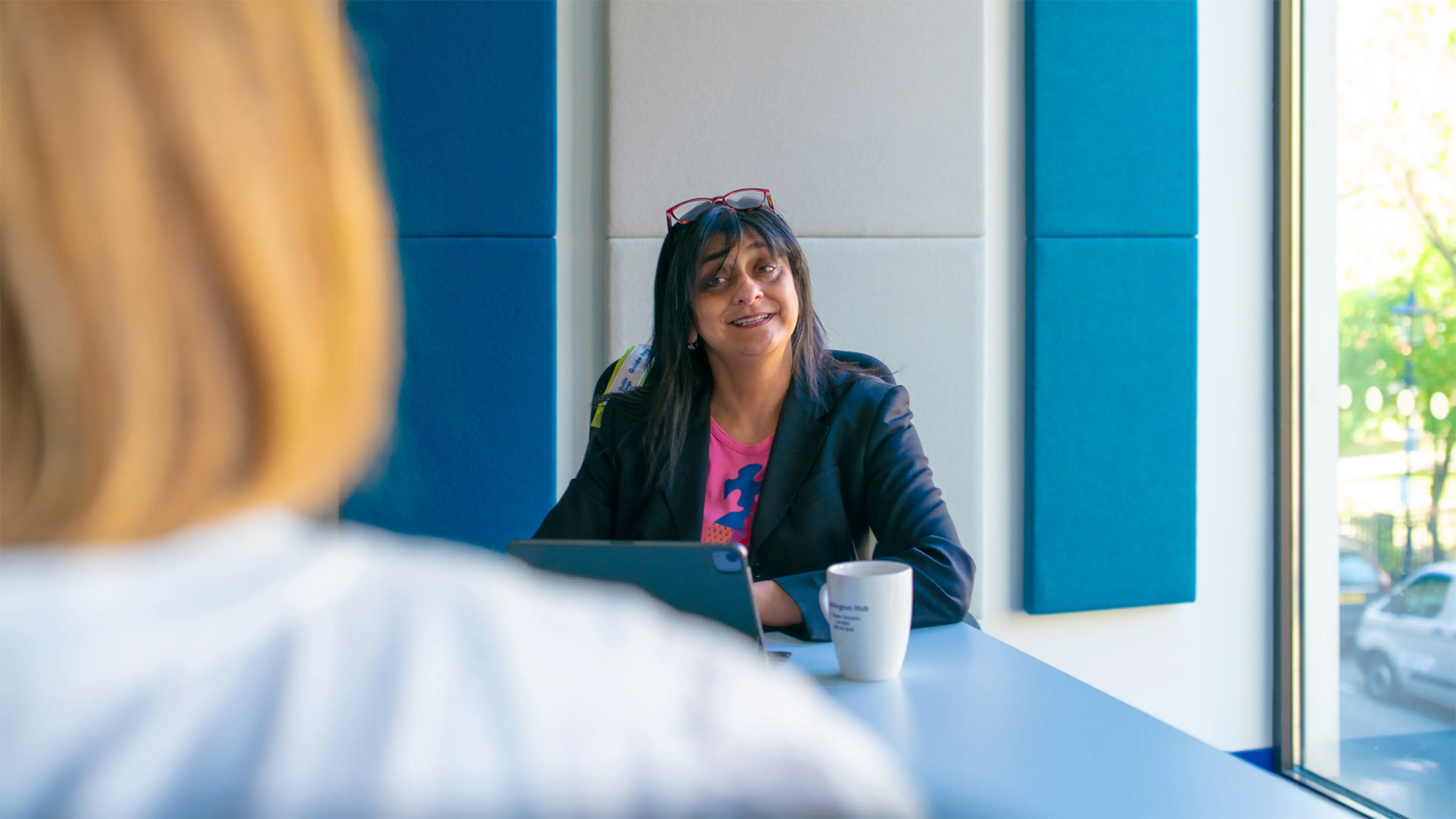 Deborah sits at a desk speaking with a colleague. Her guide dog's harness is draped on the back of her chair.