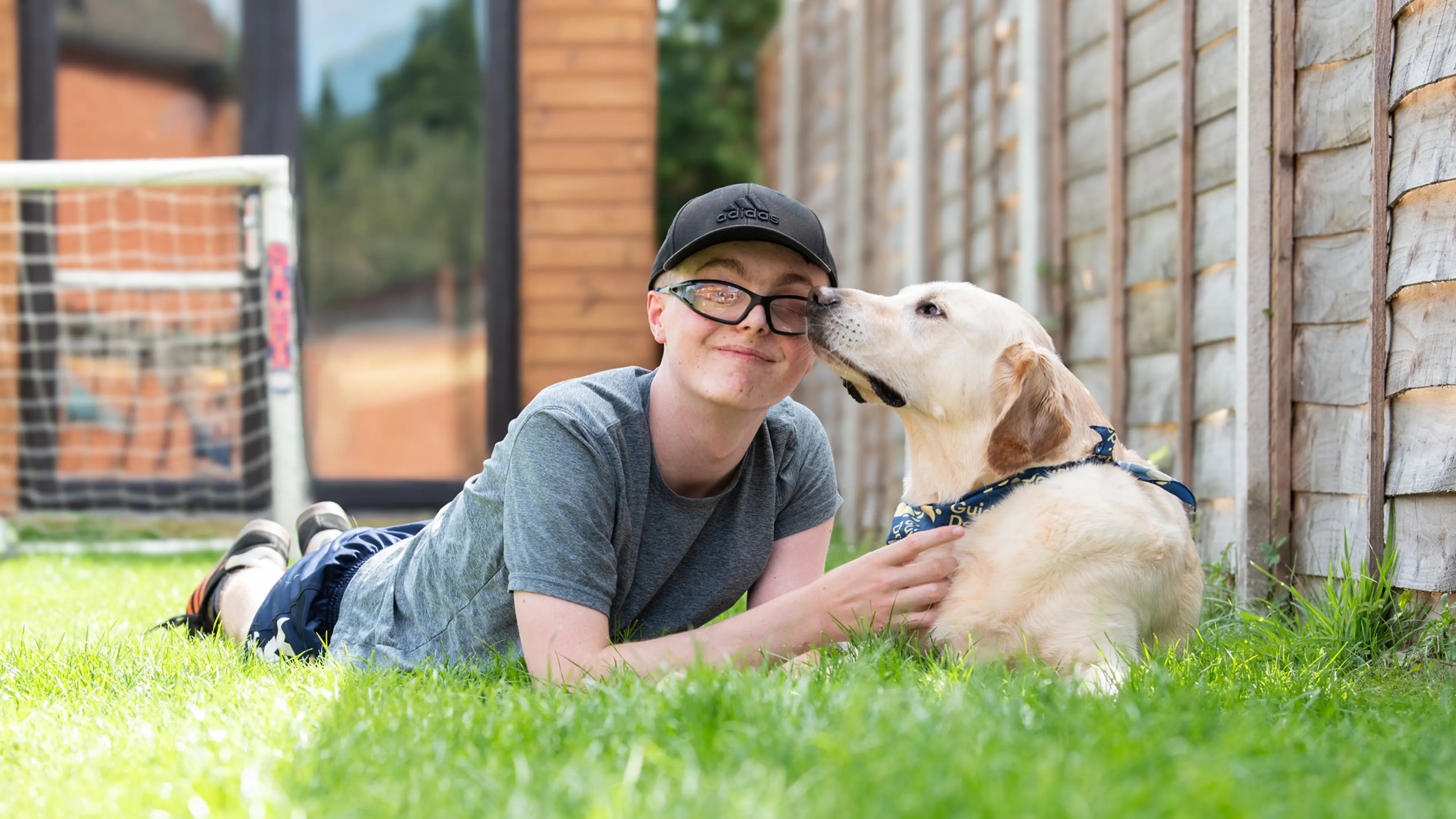 A teenager with sight loss lies on the grass in the garden with his golden retriever cross Labrador buddy dog.