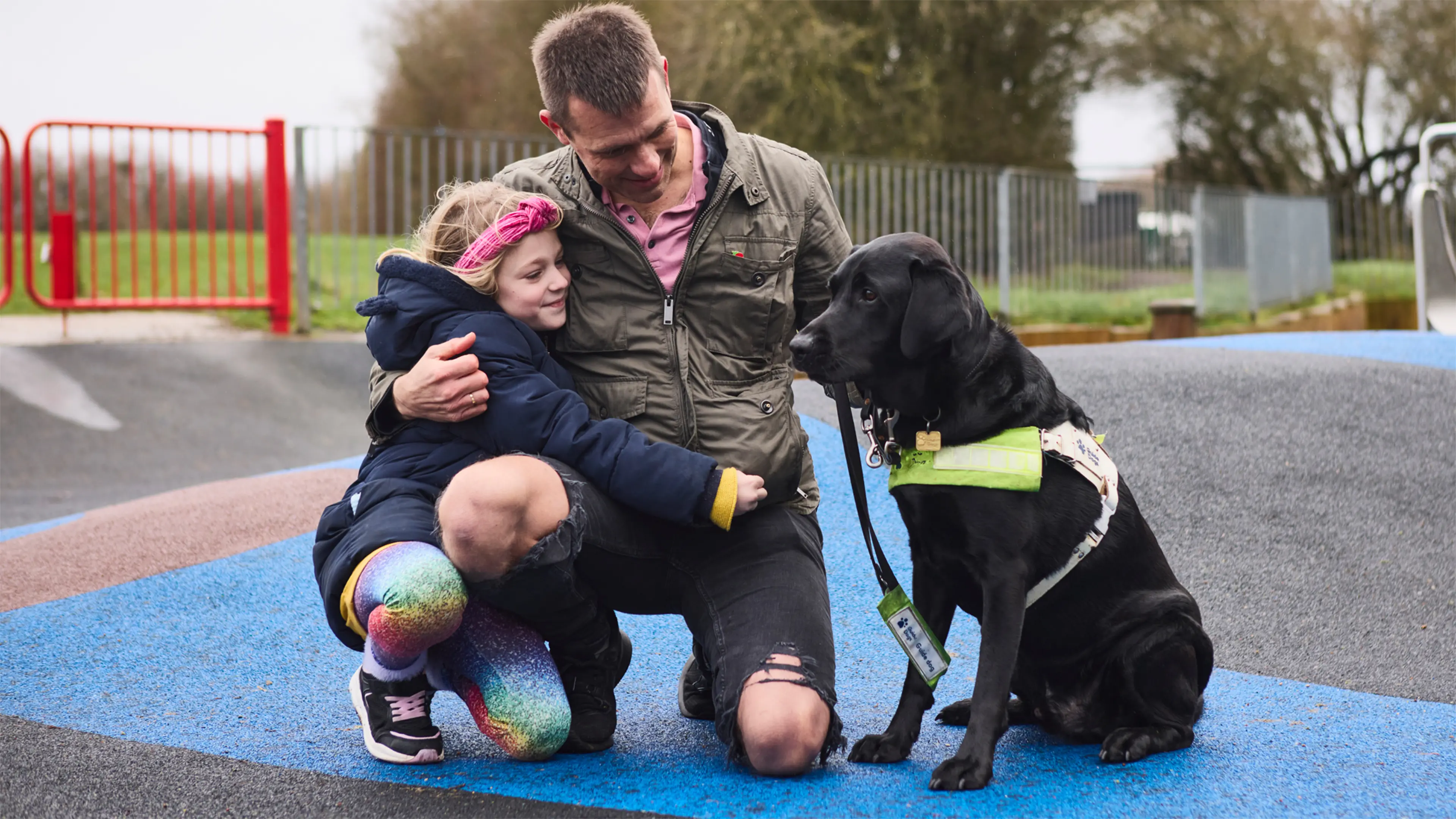 Steve and his daughter are in a playground with his black Labrador guide dog Wilf sat next to them.