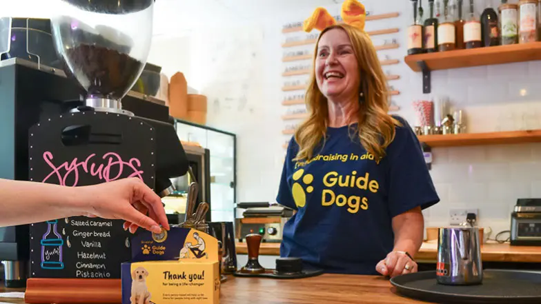 Fundraiser wearing a Guide Dogs t-shirt and dog ears smiling at a café counter whilst someone donates to a collection box