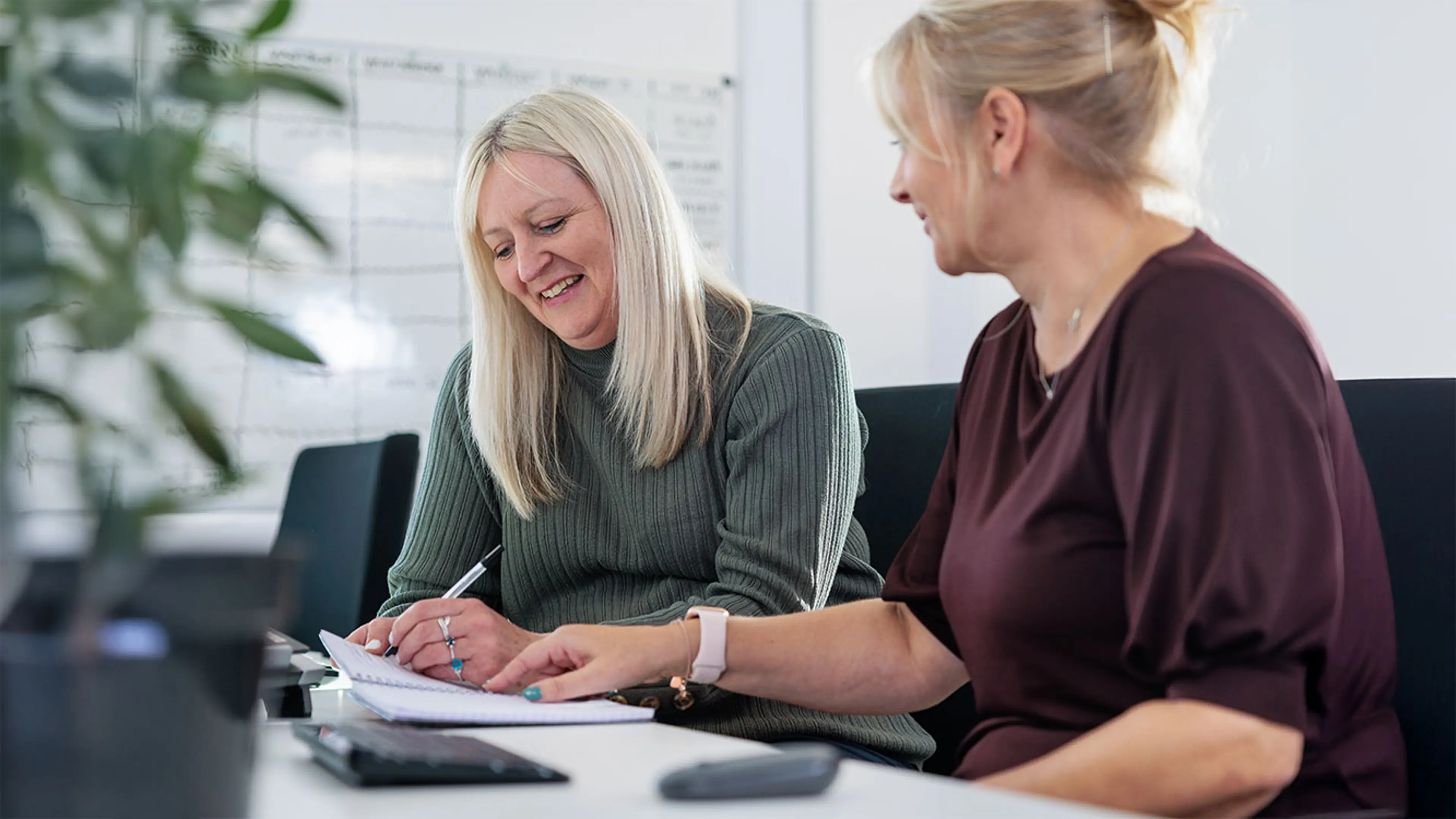 Two women are sitting at a desk, smiling while one is writing in a notebook.