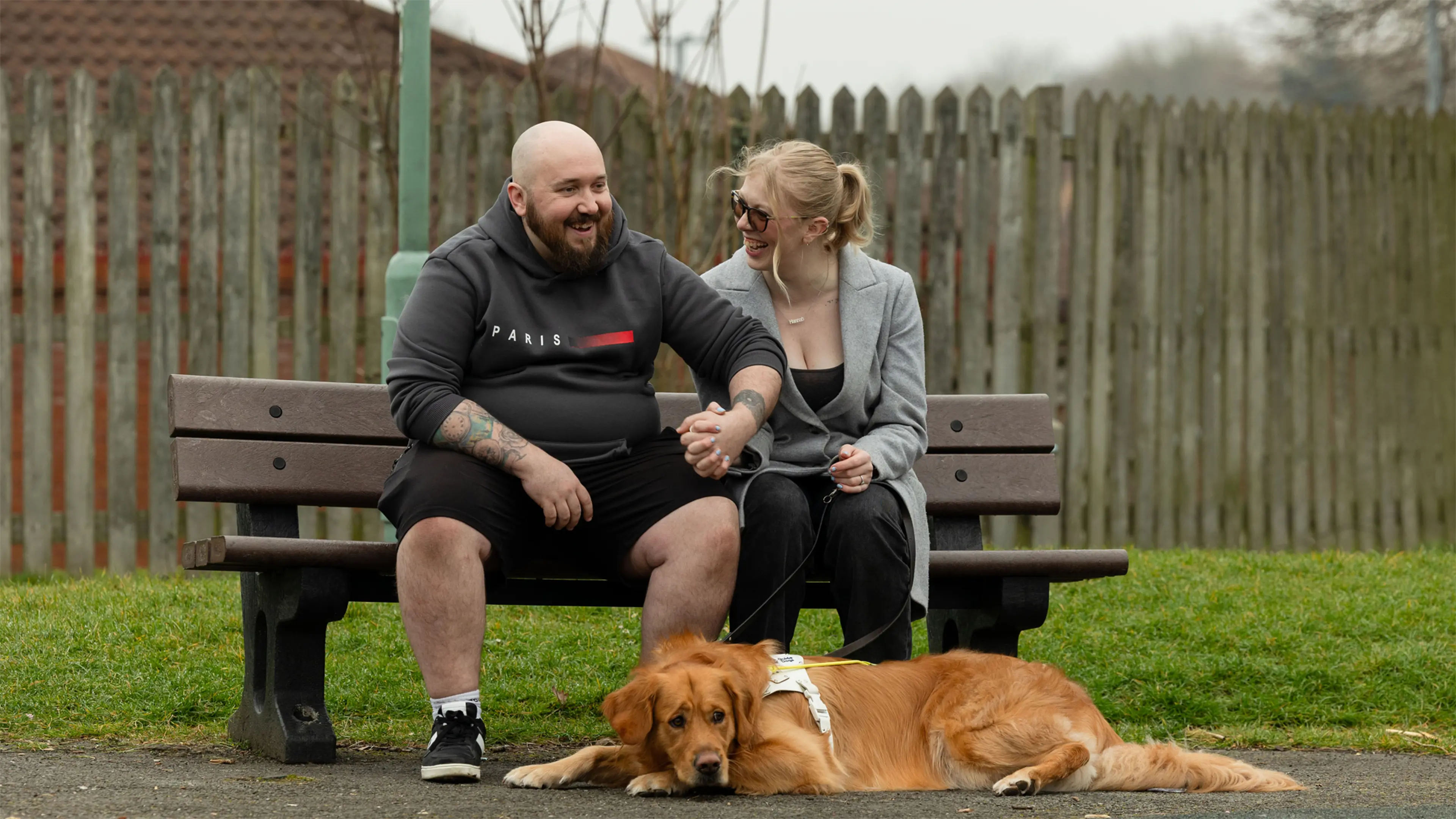 Guide dog owner Hannah sits holding hands with her fiance on a bench. Her golden retriever guide dog Morris lays on the floor at their feet.