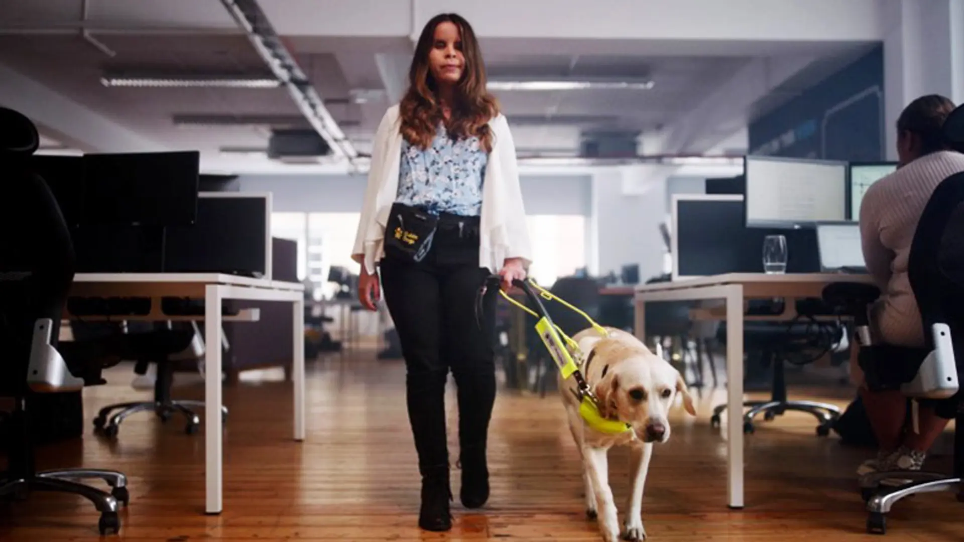 A young woman walking with her guide dog through an office.