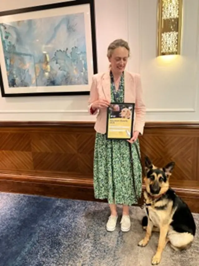 Volunteer fosterer, Cassandra, stands with her certificate at the Local Volunteer Awards with guide dog, Sable, sat next to her