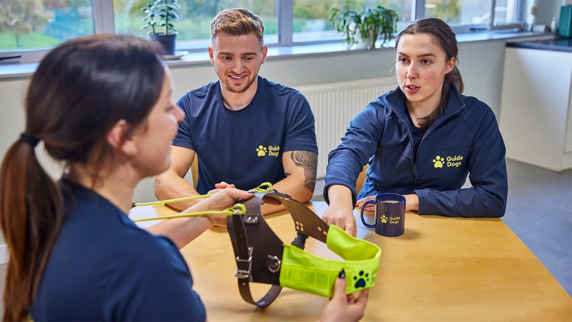 Three Guide Dog Trainers from the Guide Dogs Academy are sat at a table exploring a guide dog harness.