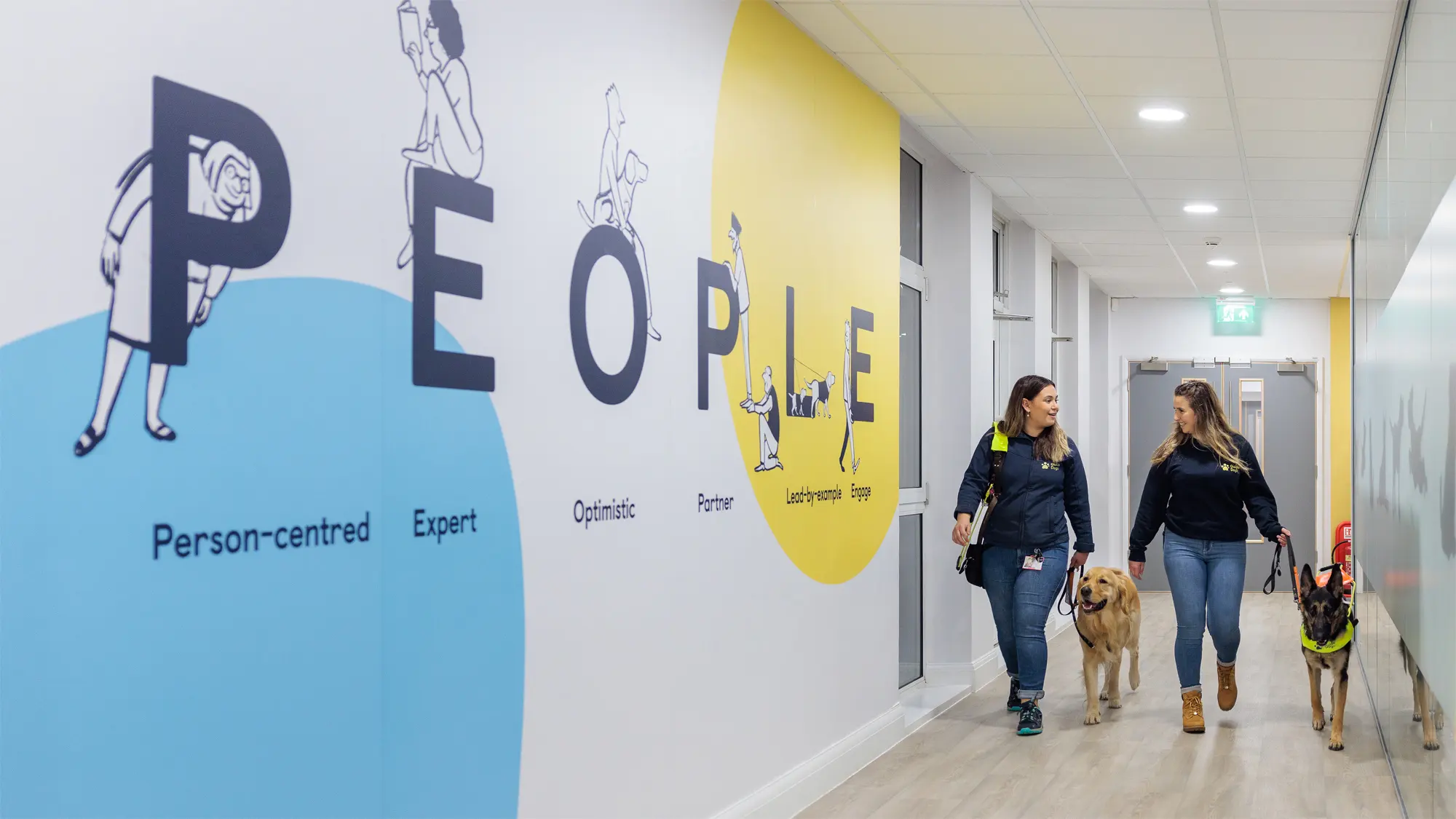 Two Guide Dog Trainers walk past a colourful wall mural that reads People, with a golden retriever and German shepherd in training.