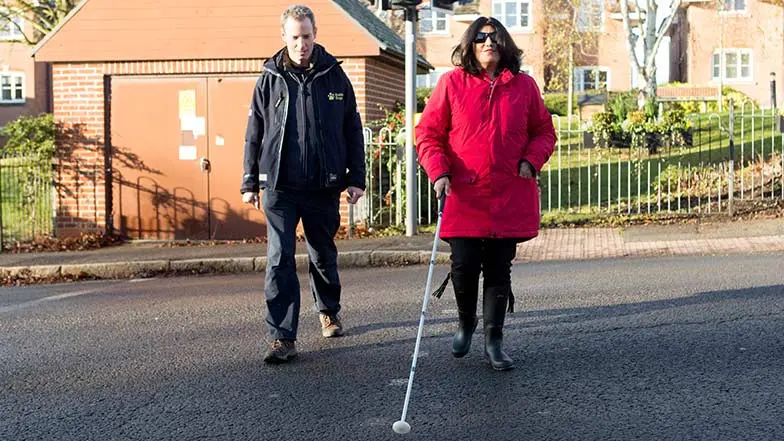 Sema is walking with a long cane across a road alongside a Vision Rehabilitation Specialist, Daniel.