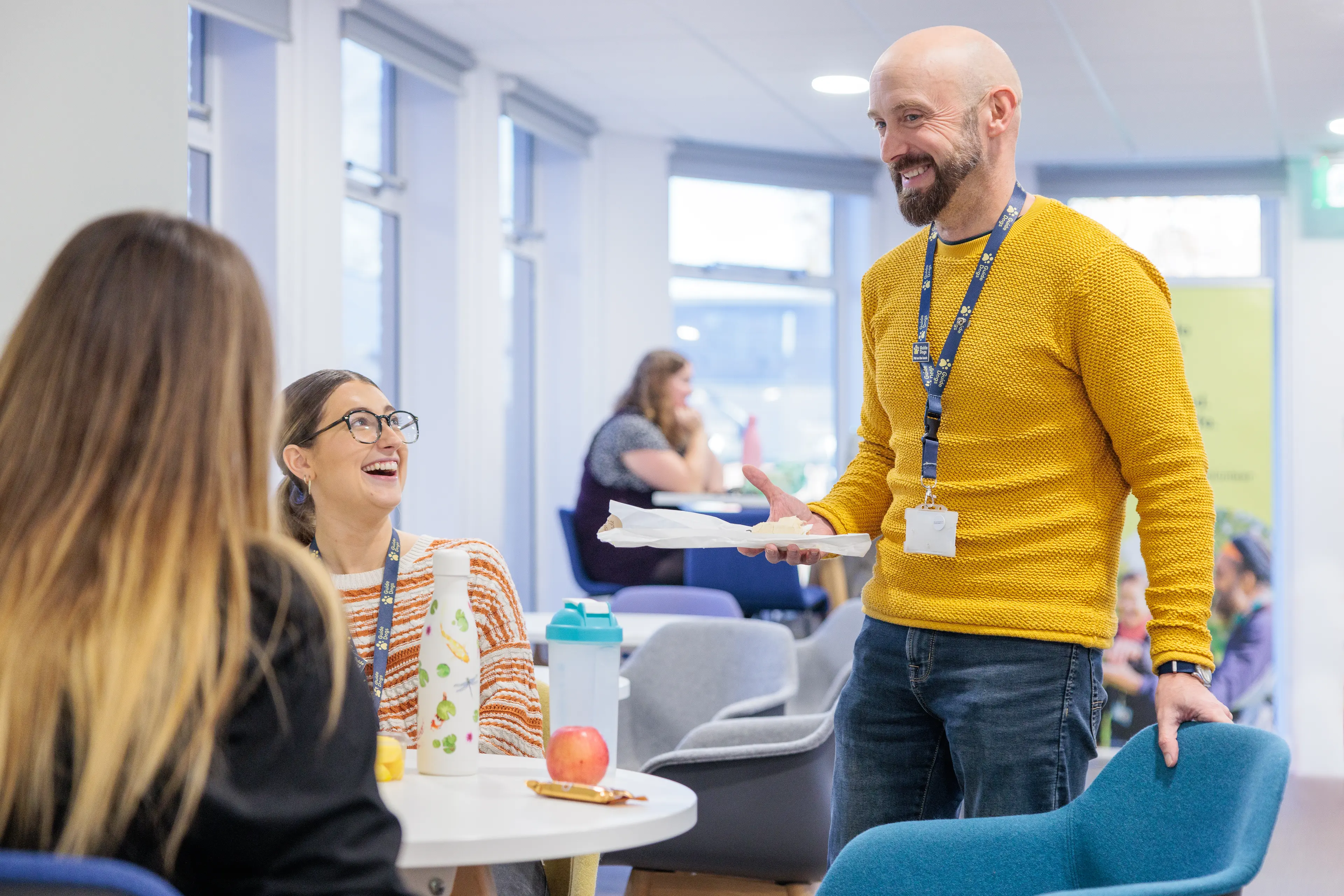 Two colleagues sit round a table laughing while another stands beside them.