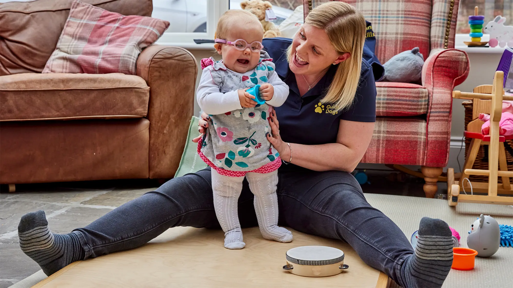 Margot, a baby who has a vision impairment, stands in the living room, supported by her Habilitation Specialist Kate. Sensory toys surround them.