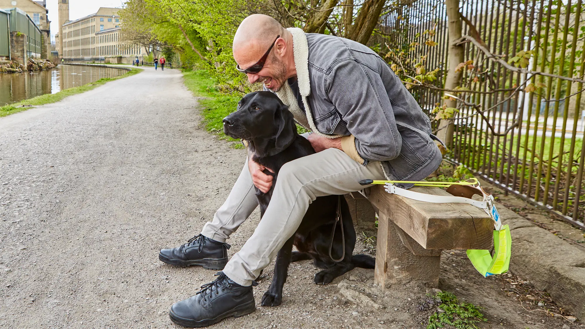 Terry, a guide dog owner, sits on a bench by a canal with his guide dog Spencer sat between his legs.
