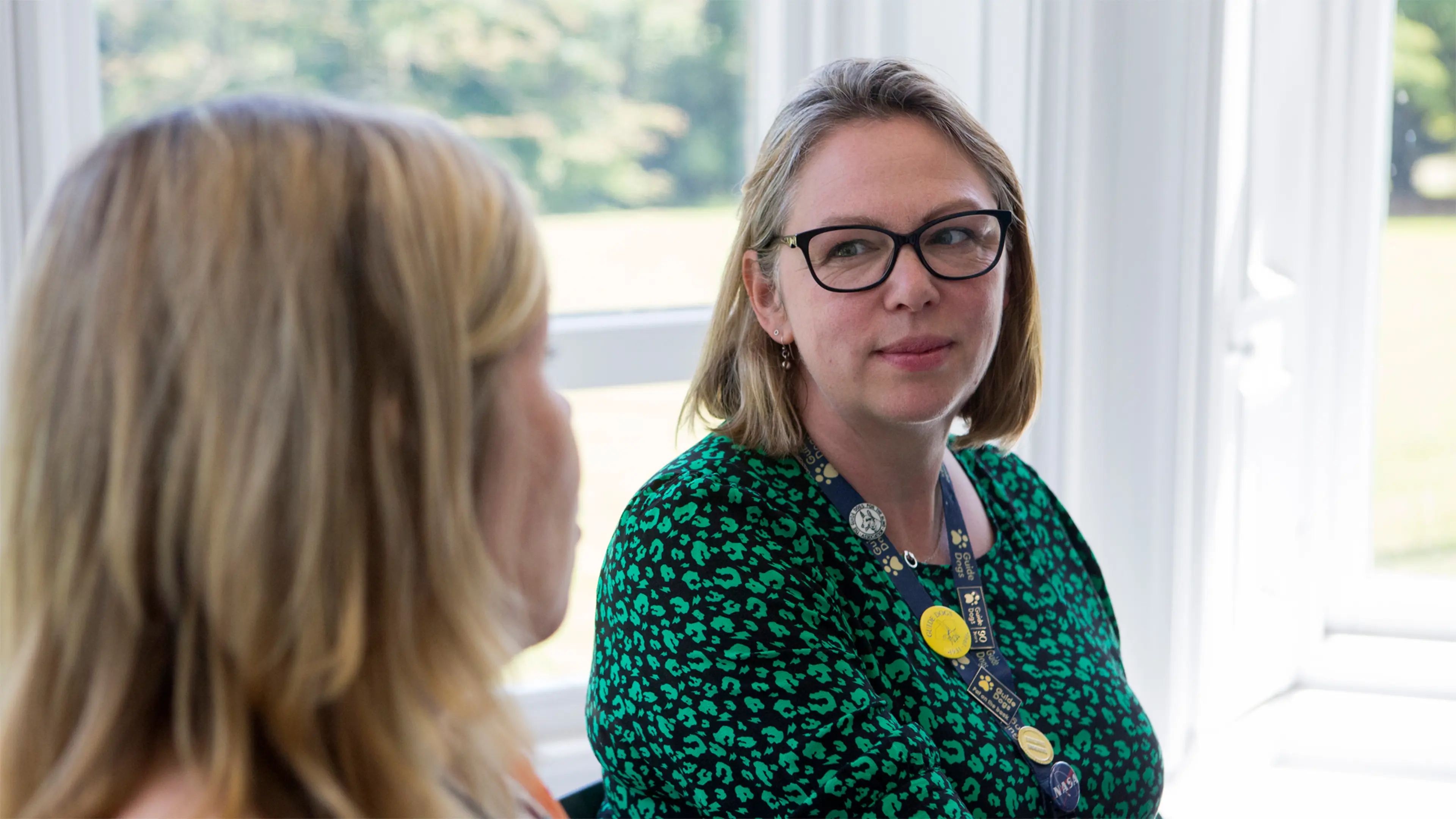 Two Guide Dogs colleagues sit by a window in the Reading office talking. One of them wears a Guide Dogs lanyard.