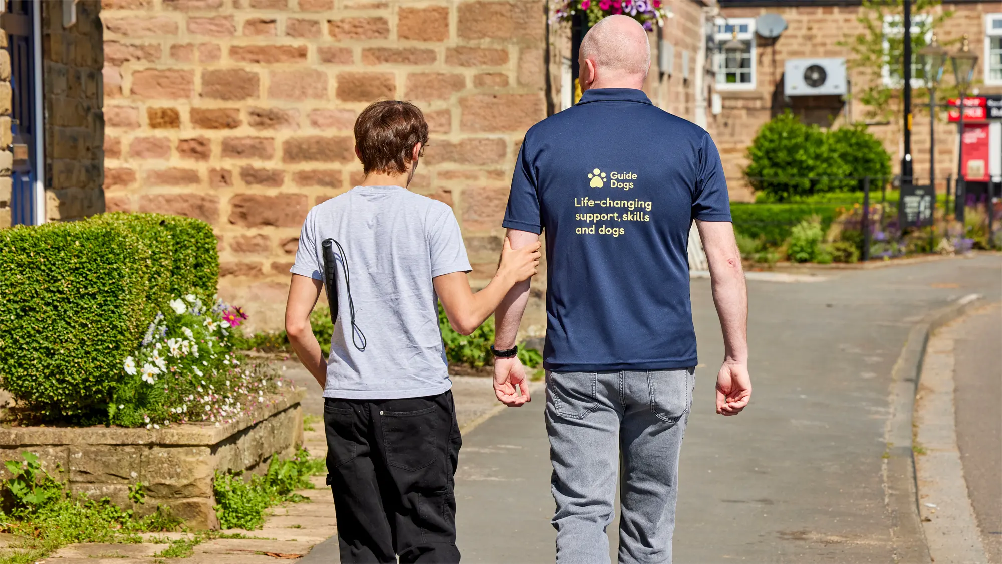 A teenager with sight loss is guided along the pavement by his Guide Dogs Habilitation Specialist.