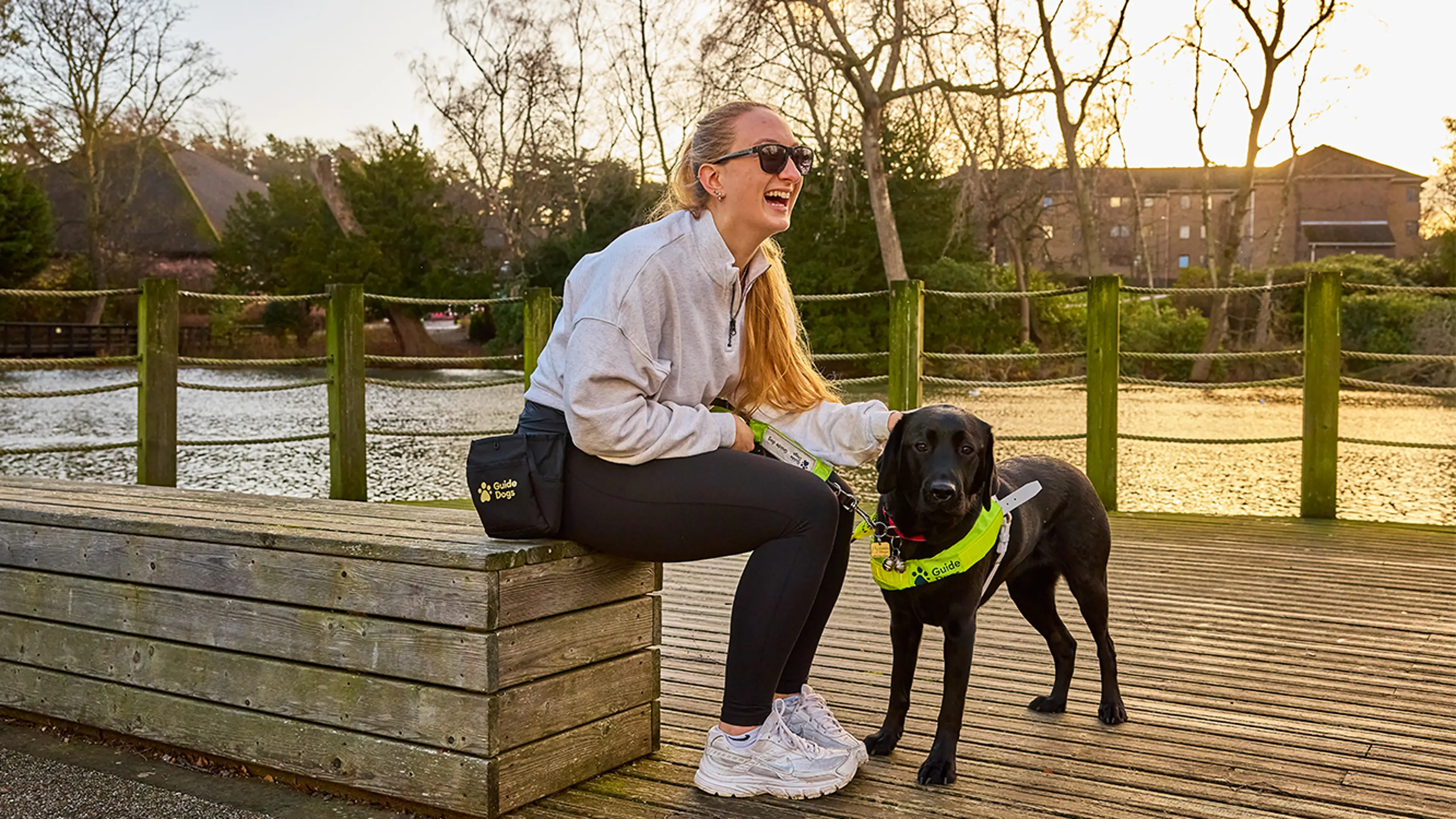 Sam sits on a riverside bench, laughing as she strokes her guide dog, Helga.