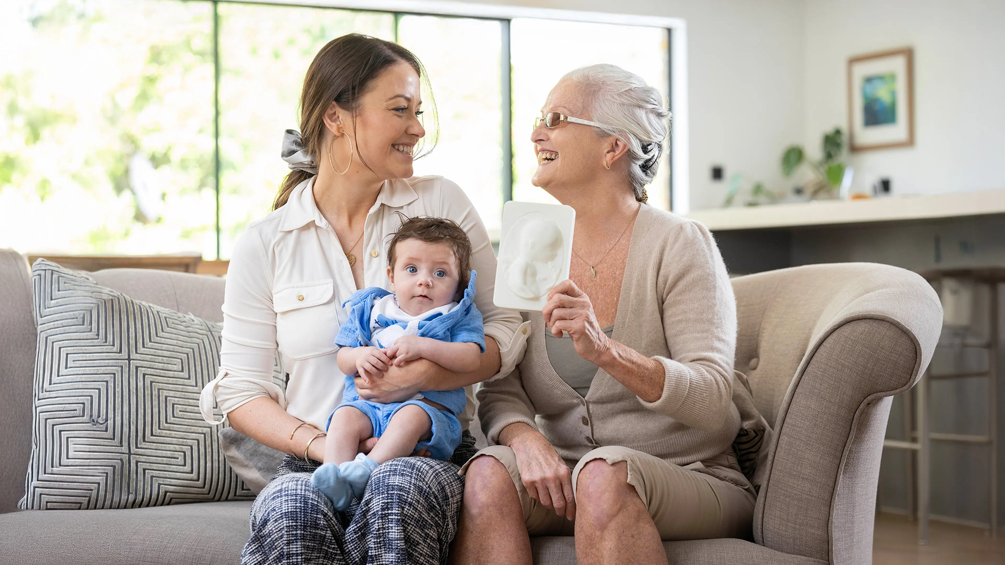 Debbie sits with her daughter Jess and baby grandson Freddie on the sofa. Debbie and Jess are smiling at each other as Debbie holds up her First Hello tactile sonogram of her grandson up to the camera.