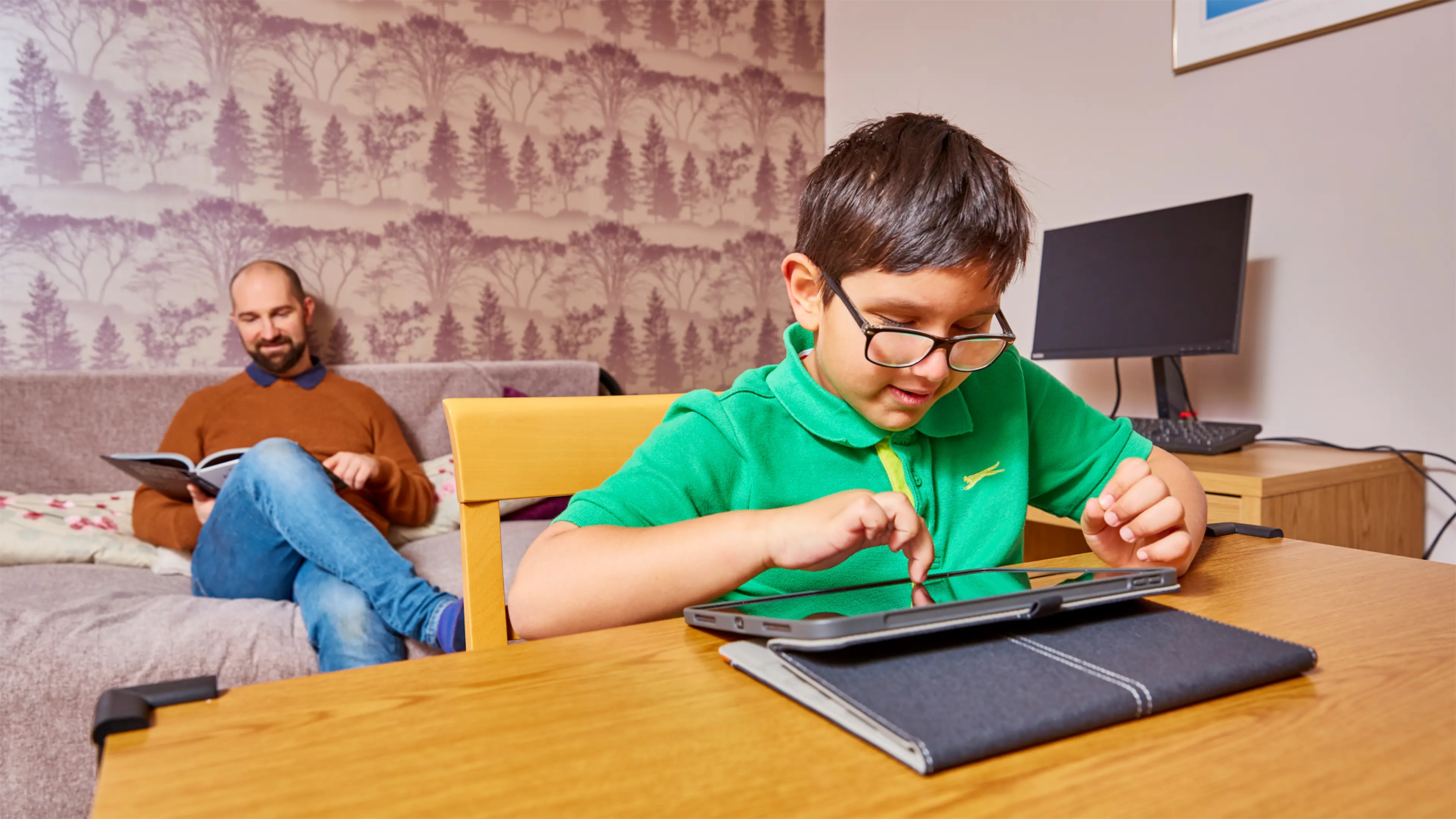 Teddy, a child who has a vision impairment, sits playing independently on a tablet at the table. His dad is sitting on the sofa in the background.
