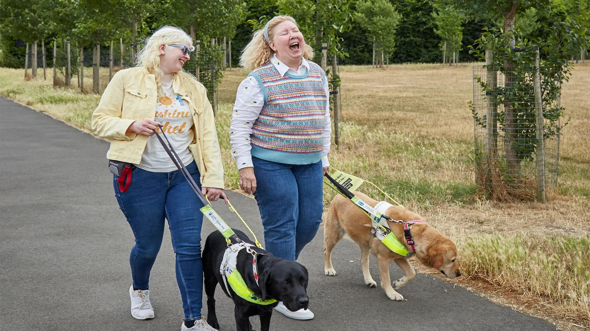 Guide dog owners Karishma and Jane laugh as they walk down a tree-lined path with their guide dogs. 