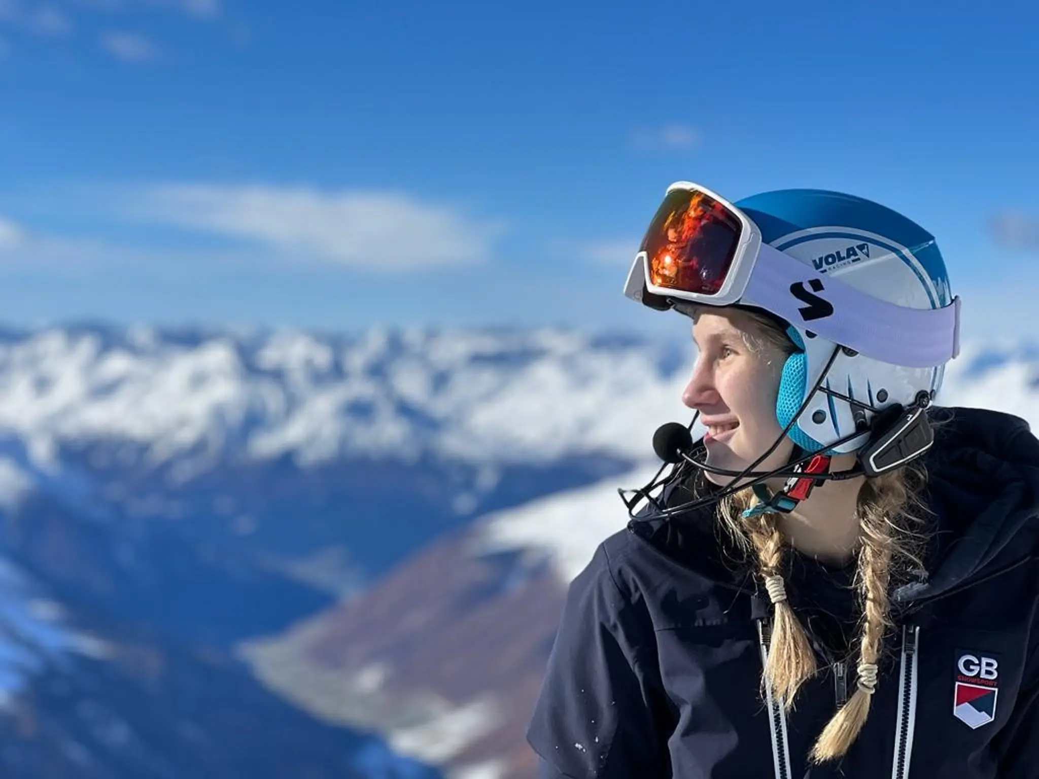 Hester, a guide dog owner, smiles as she sits wearing her ski goggles and a helmet. Behind her are snow covered mountains. 