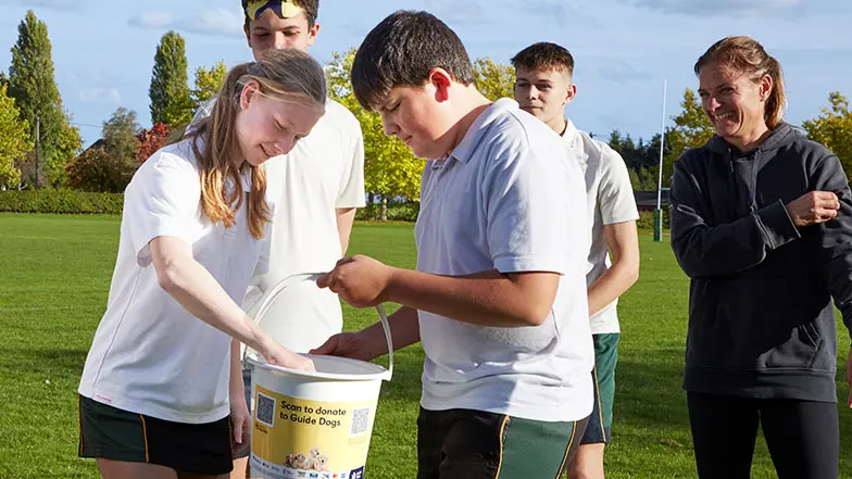 School children in PE kit paying in money into a Guide Dogs collection bucket.