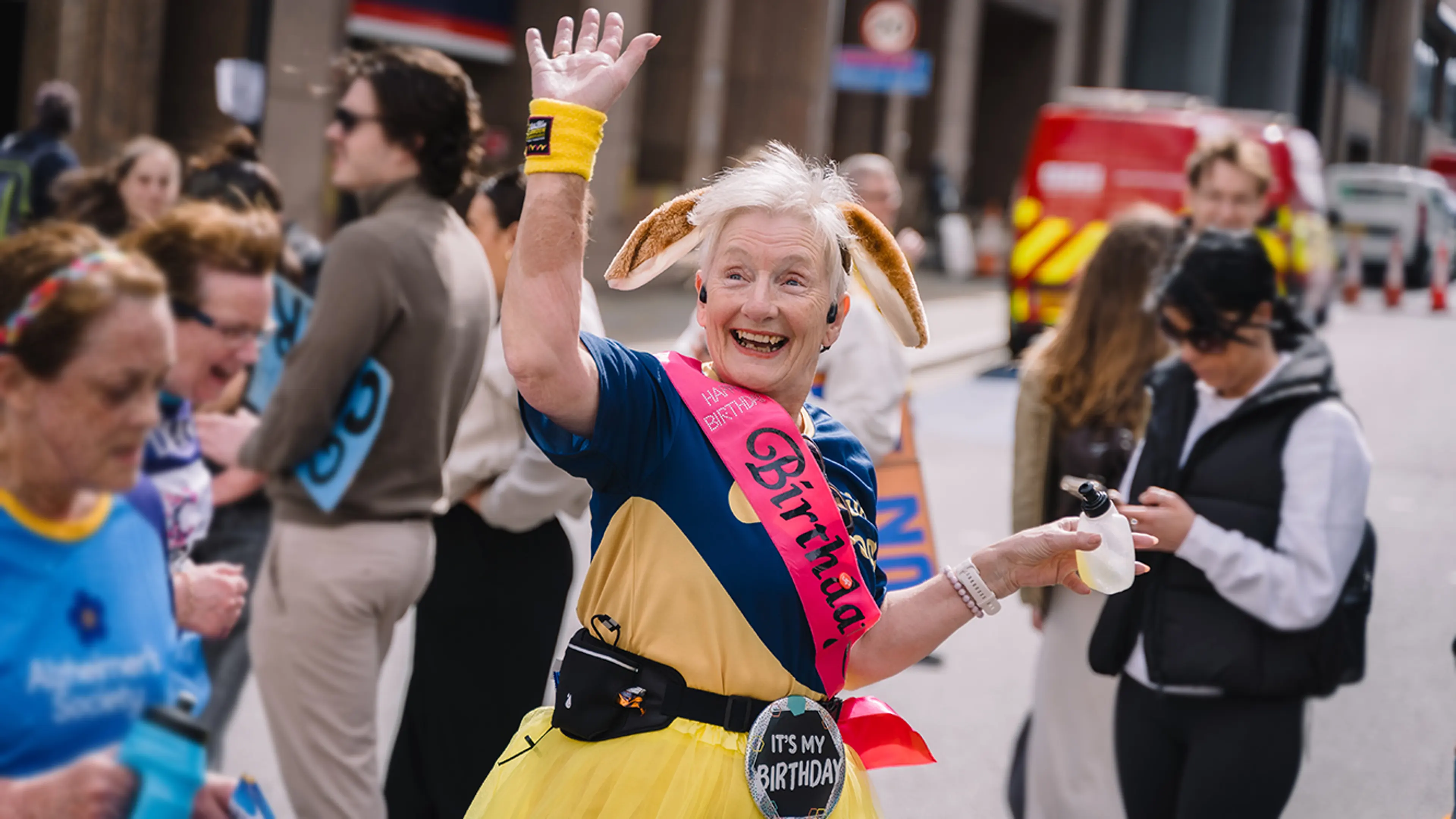 A lady wearing a birthday sash on top of her Guide Dogs marathon Tshirt waving at her supporters as she runs the London Landmarks Half Marathon.
