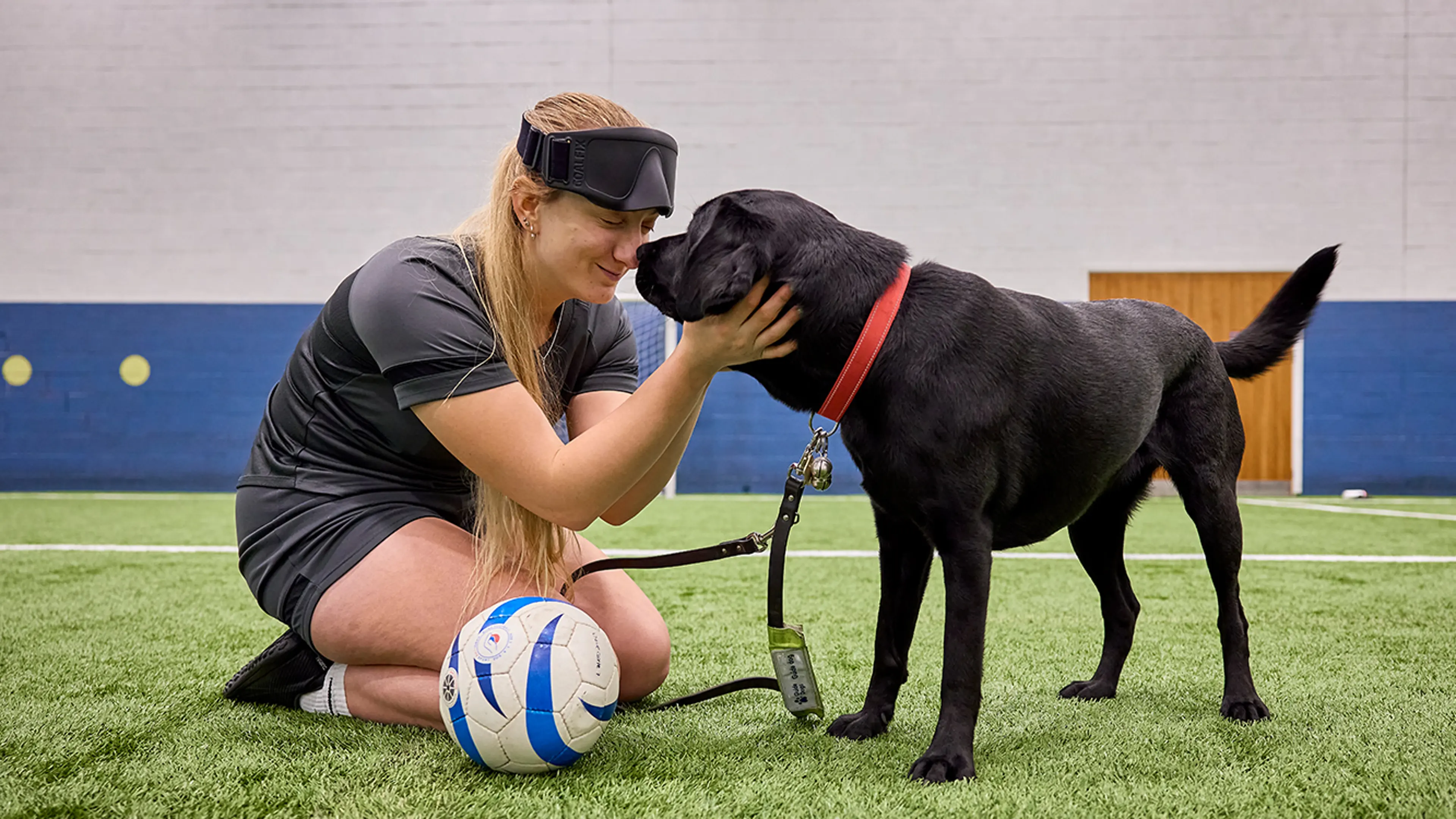Sam and her guide dog, Helga, touch noses in a gym while Sam smiles warmly