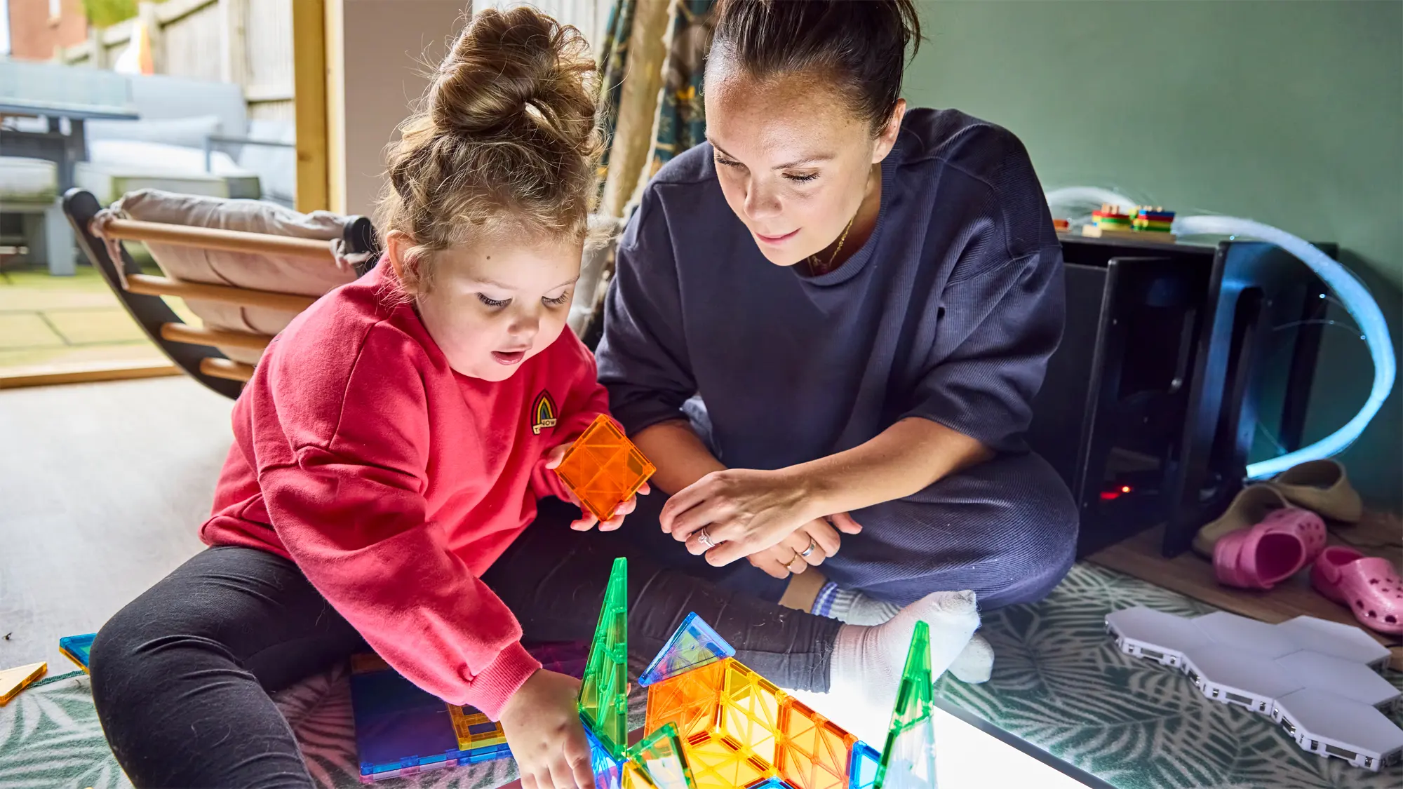 A young girl with vision impairment and her mum play with some colourful building blocks on a bright white mat.